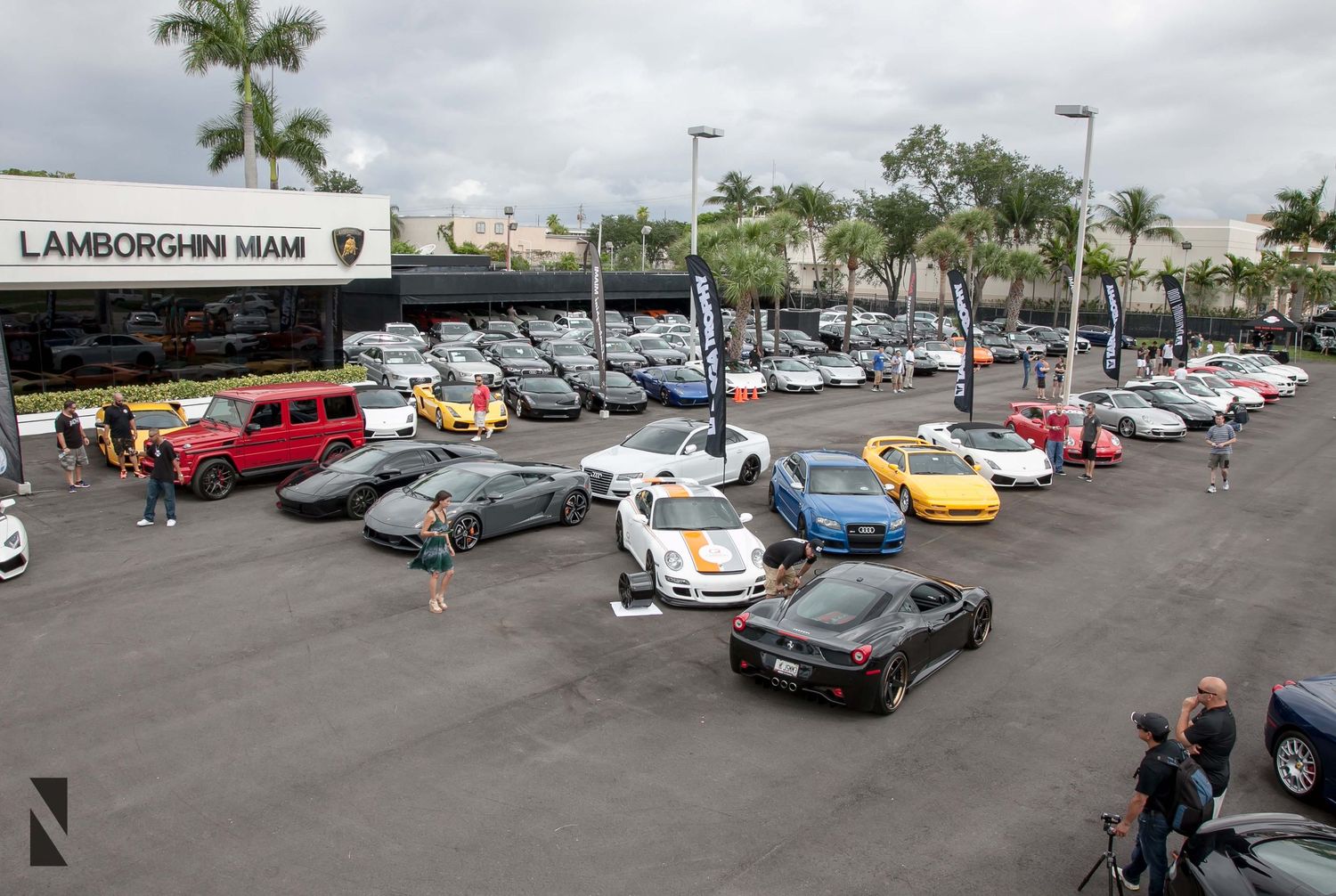 Row of luxury and exotic cars parked outside a Lamborghini dealership under cloudy skies.