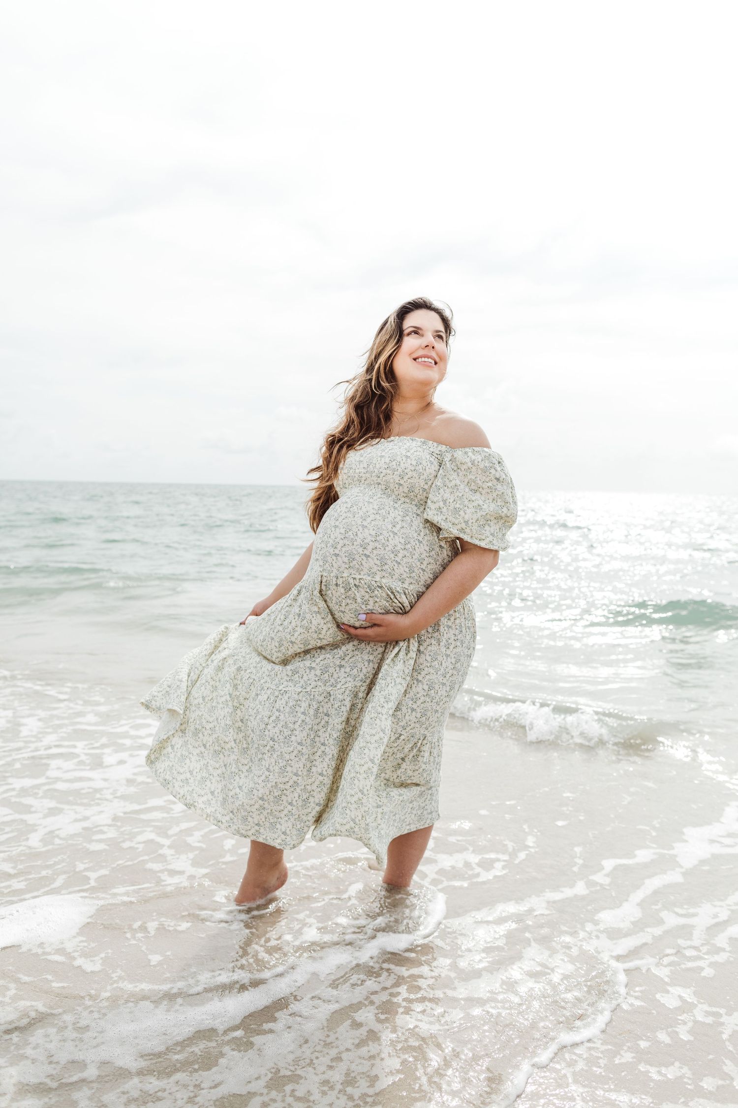 A person in a floral off-shoulder dress stands in shallow ocean waves at the beach while smiling and holding their belly.