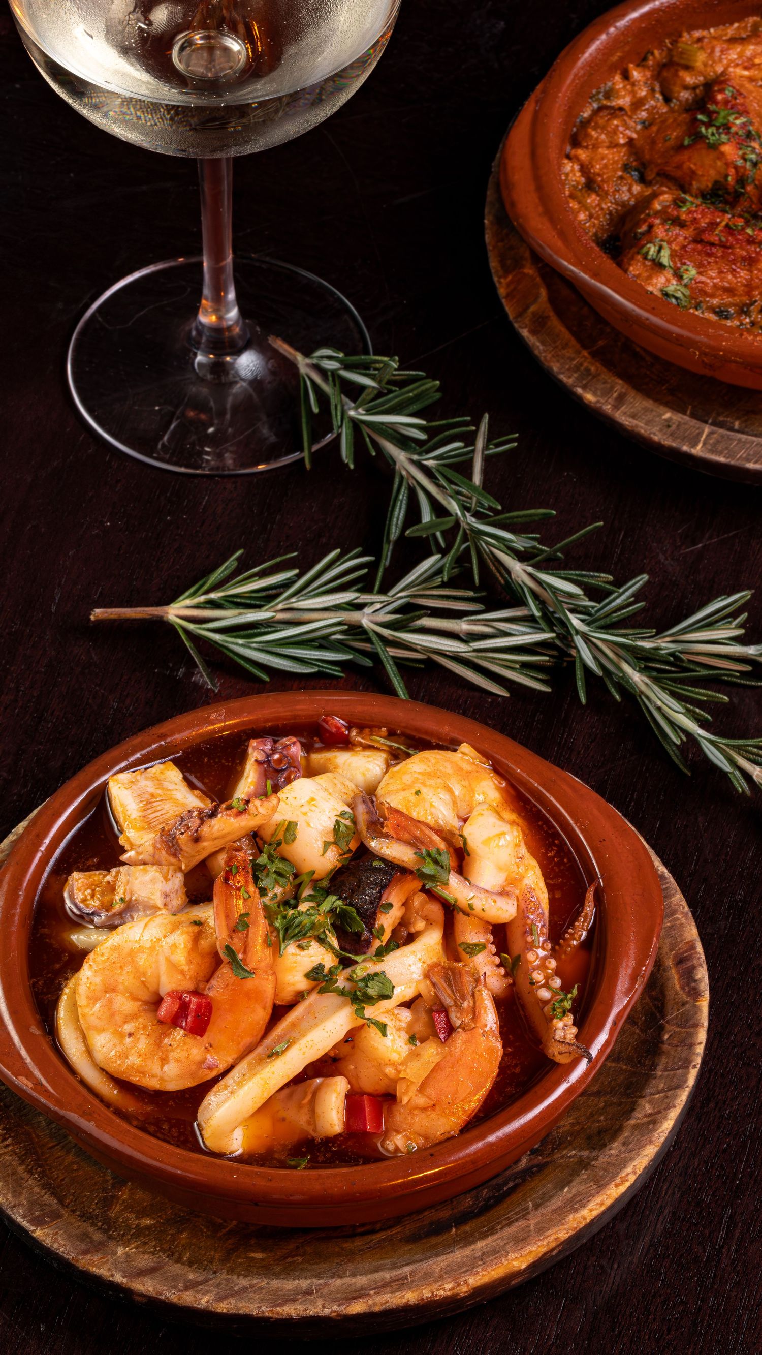 Seafood stew with octopus and shrimp served in terracotta dish with fresh rosemary and wine glass on dark wooden table.