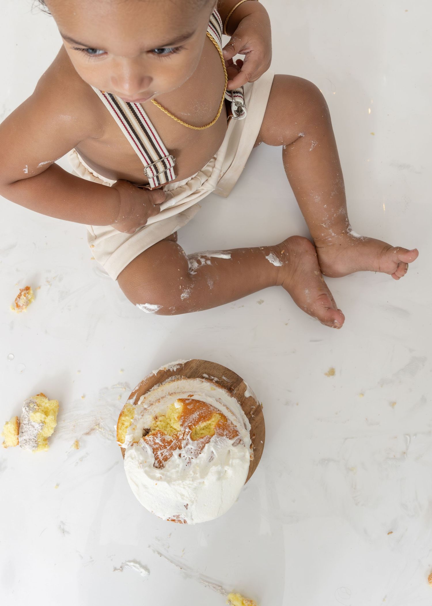 A baby in a white onesie sits on the floor while enjoying a cake smash photoshoot with a white frosted cake.
