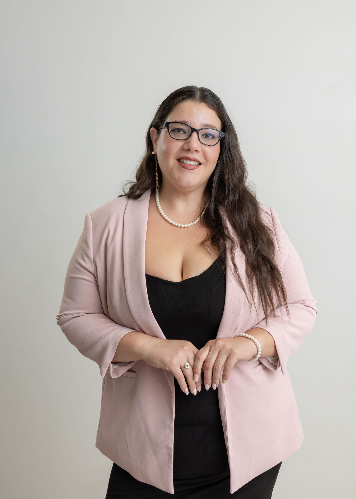 Professional portrait showing a person in a pink blazer and black top against a light background, wearing glasses and pearls.