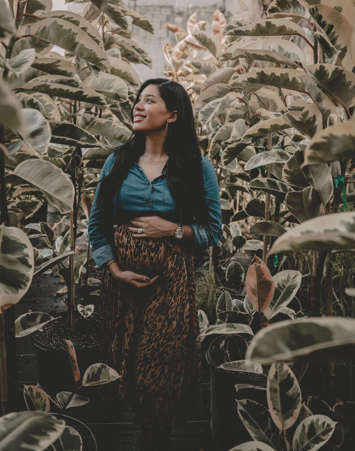 A woman in a leopard print skirt stands among banana leaves while cradling her pregnant belly.