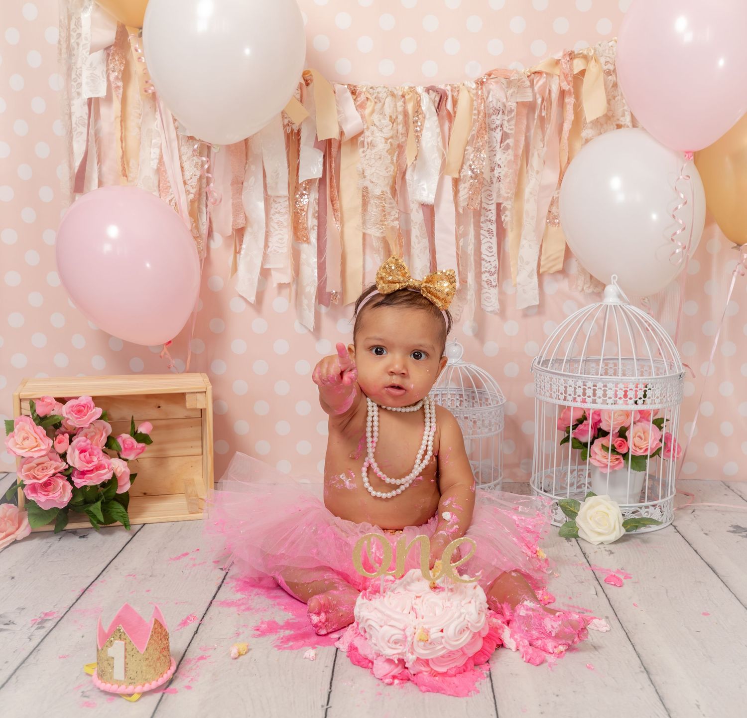 Pink and white first birthday cake smash photo setup with tutu, balloons, floral decor and birdcage on wood floor.