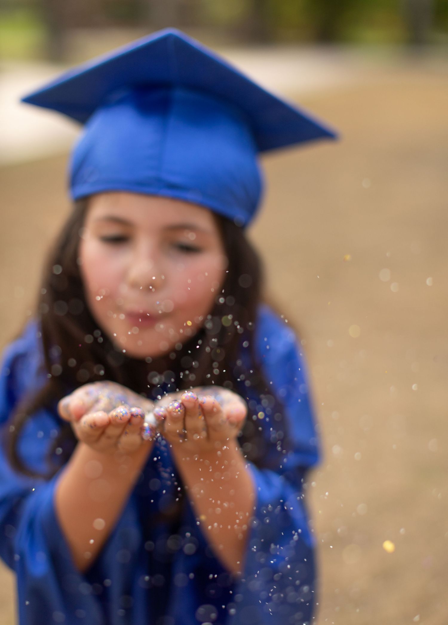 Graduate in blue cap and gown blows glitter from cupped hands in celebratory graduation portrait.