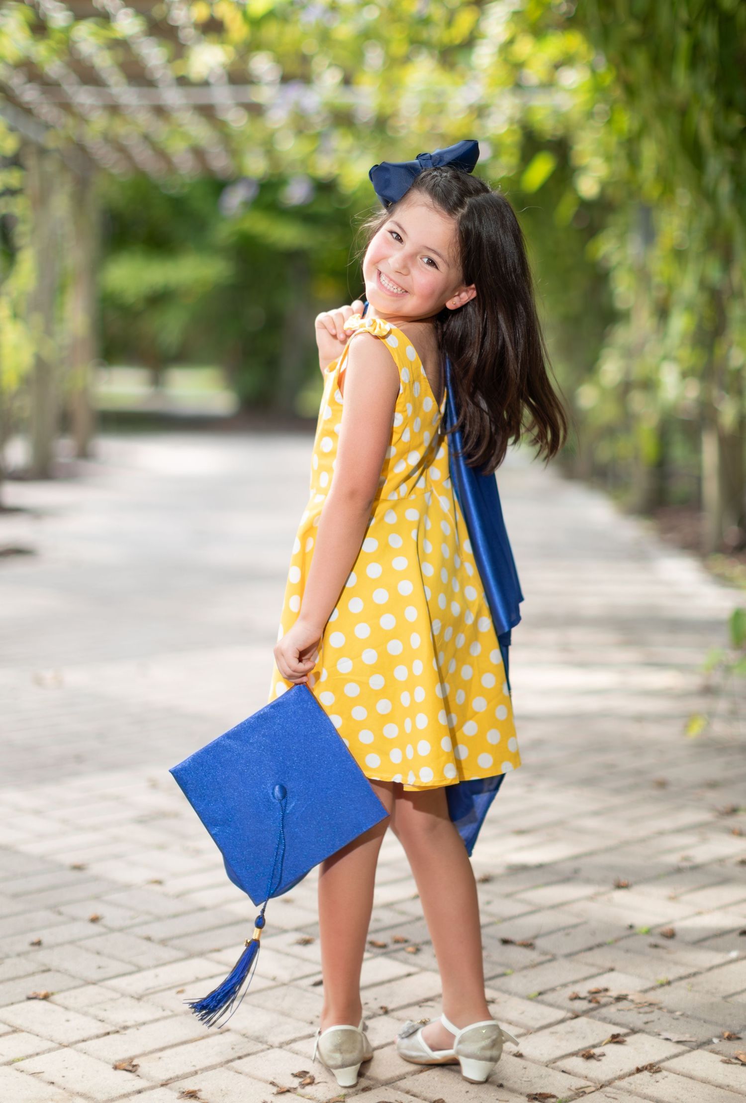 Young student in yellow polka dot dress with blur graduation cap stands on garden path on graduation day.