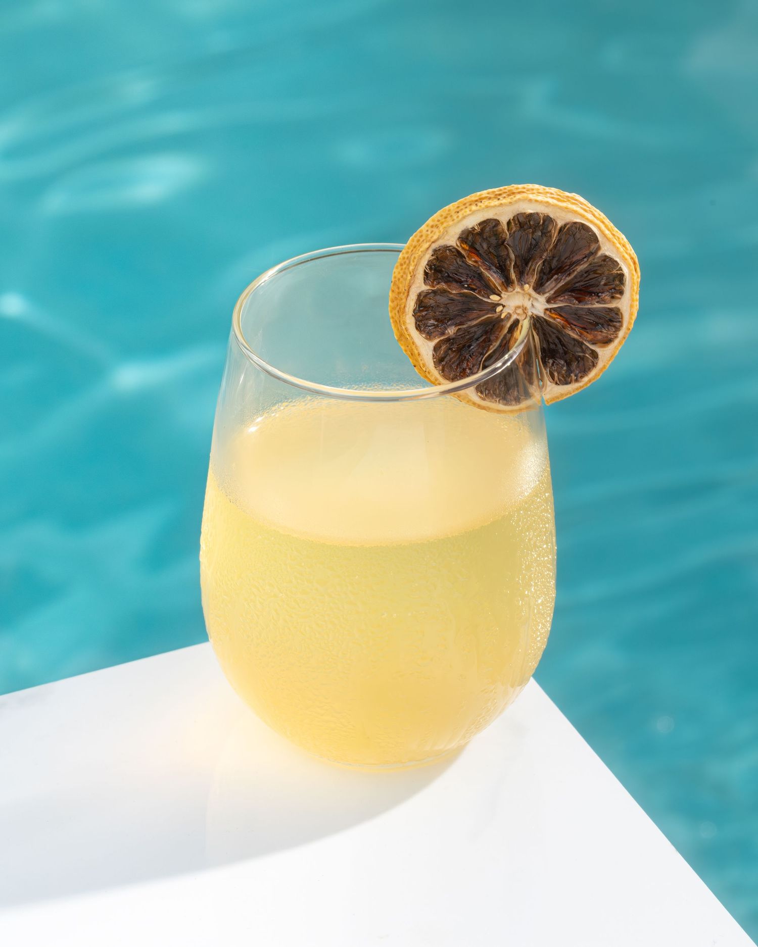 A refreshing citrus cocktail with a blood orange garnish sits by a pool with turquoise water in the background.