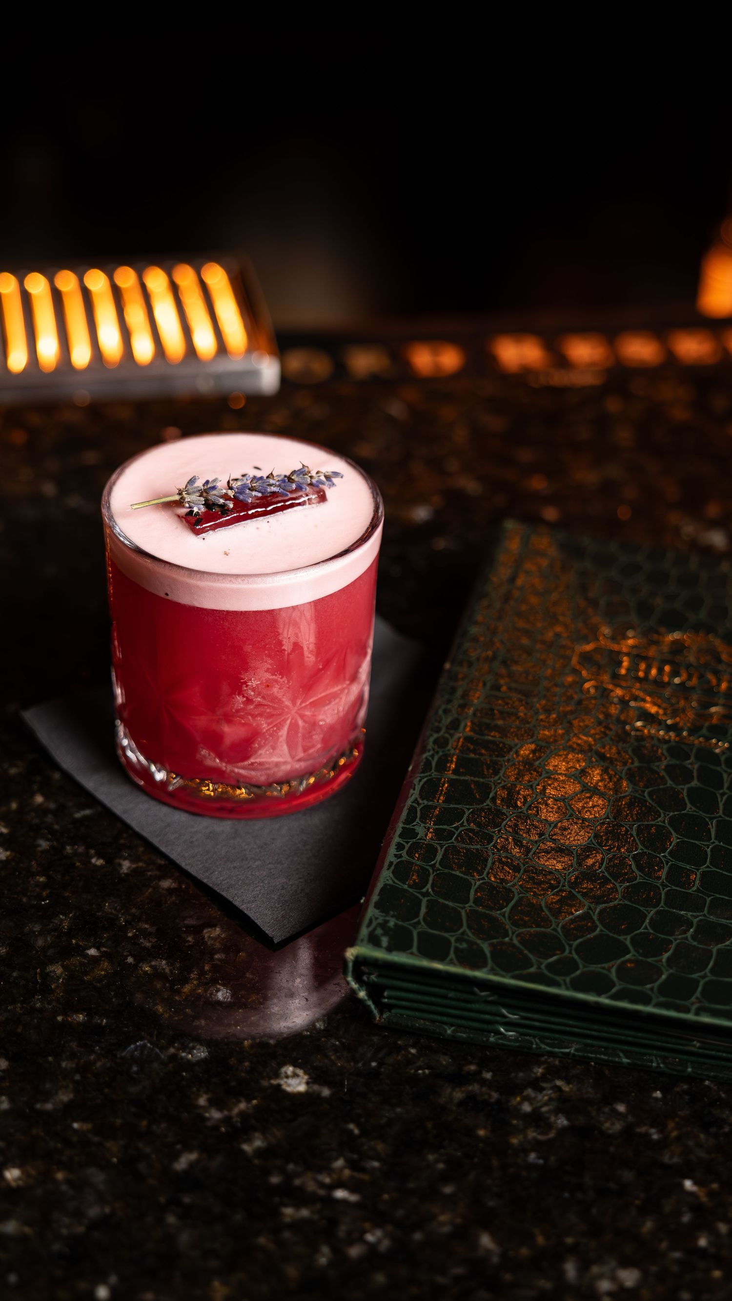 Red craft cocktail with pink foam and garnish served on a dark surface next to a green book with ambient lighting.