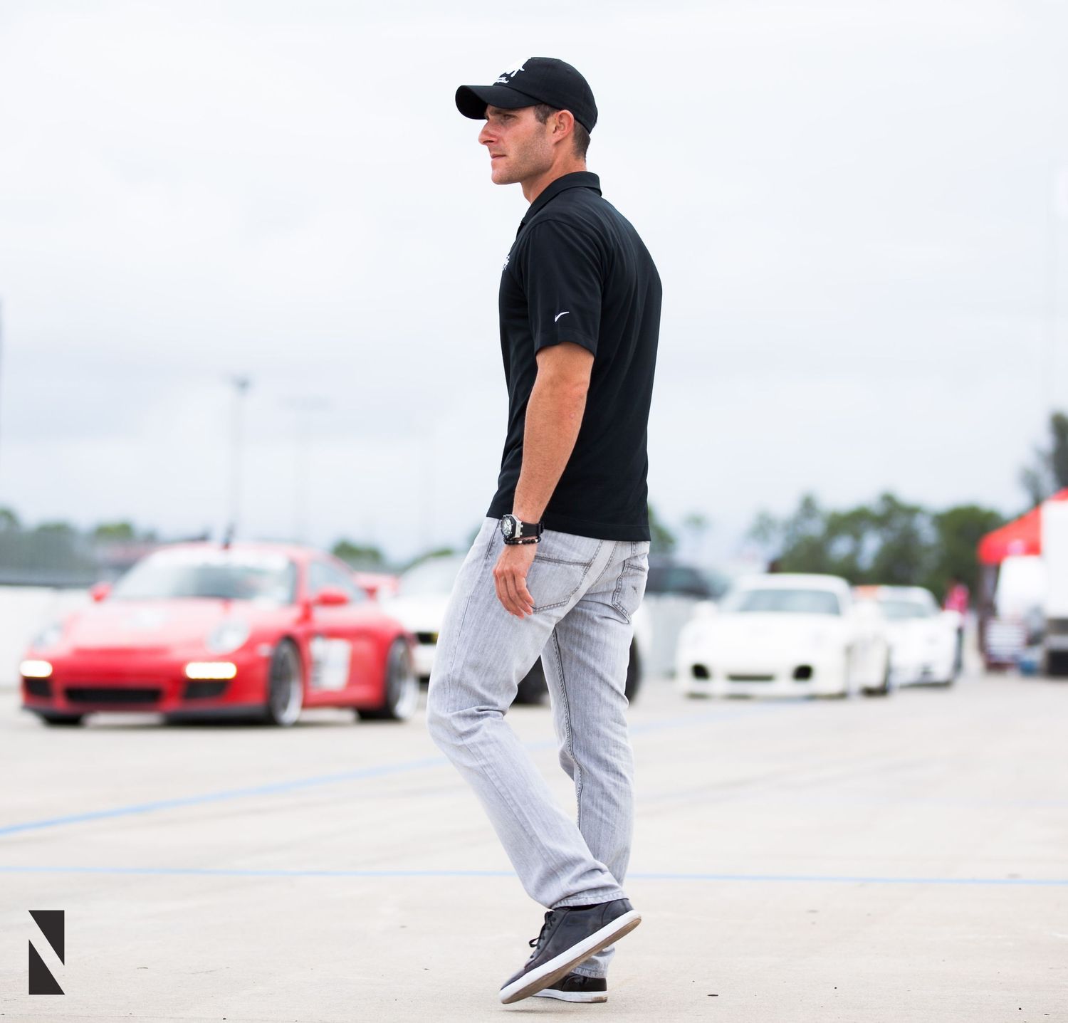 A person in black shirt and light jeans stands in a parking lot with sports cars visible in the background.