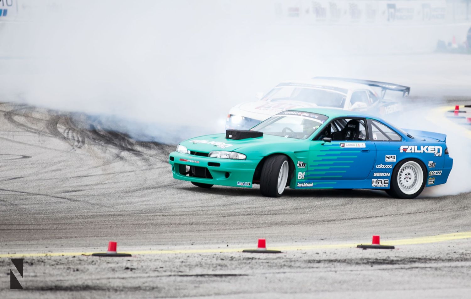 A turquoise and blue drift car slides sideways around a track leaving tire marks near orange traffic cones.