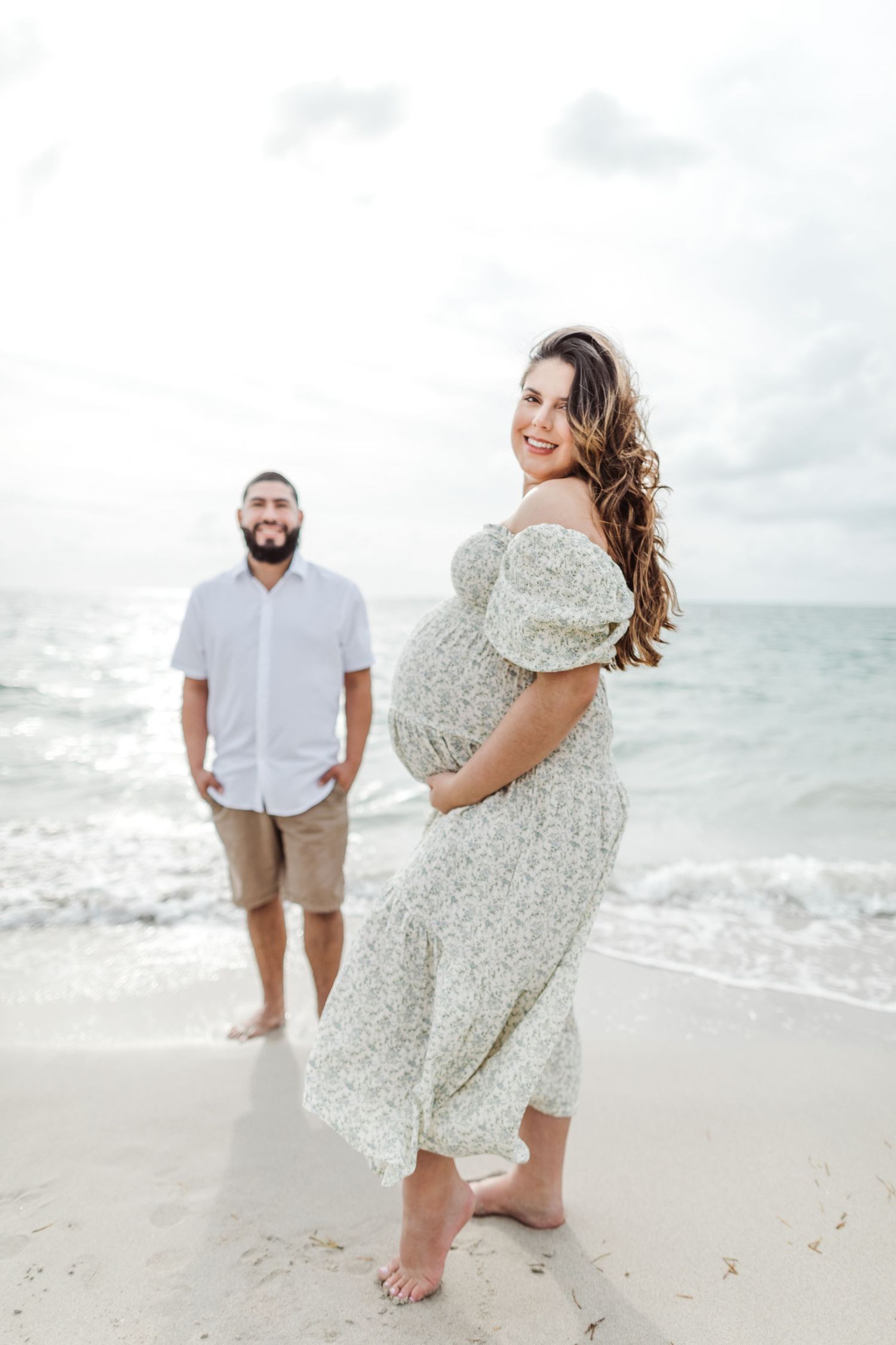 A pregnant woman in a flowy white dress smiles joyfully on the beach with her partner standing in the background.