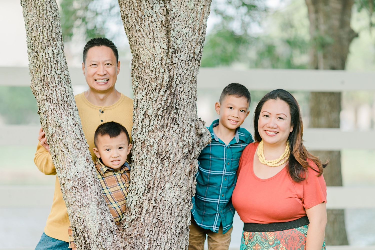 Happy family of four posing together by a tree trunk outdoors in casual clothing.