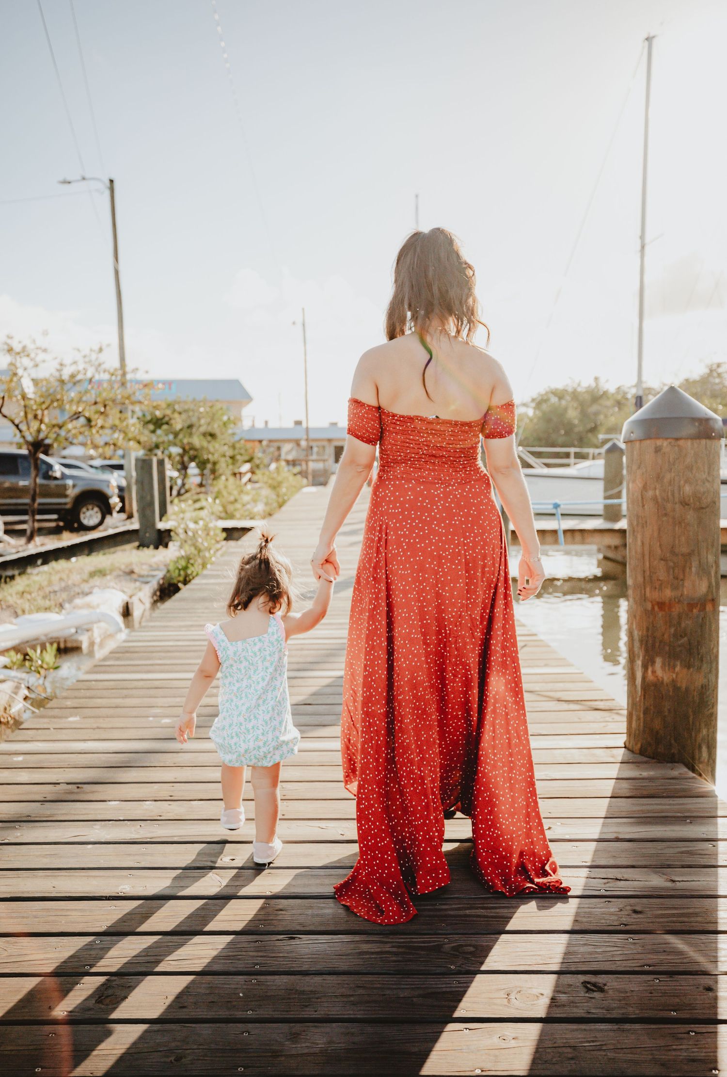 Woman in red dress holding hands with small child walking on wooden dock at sunset.