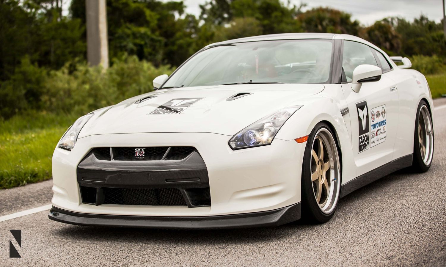 White Nissan GT-R with aftermarket wheels and body kit on a rural road.