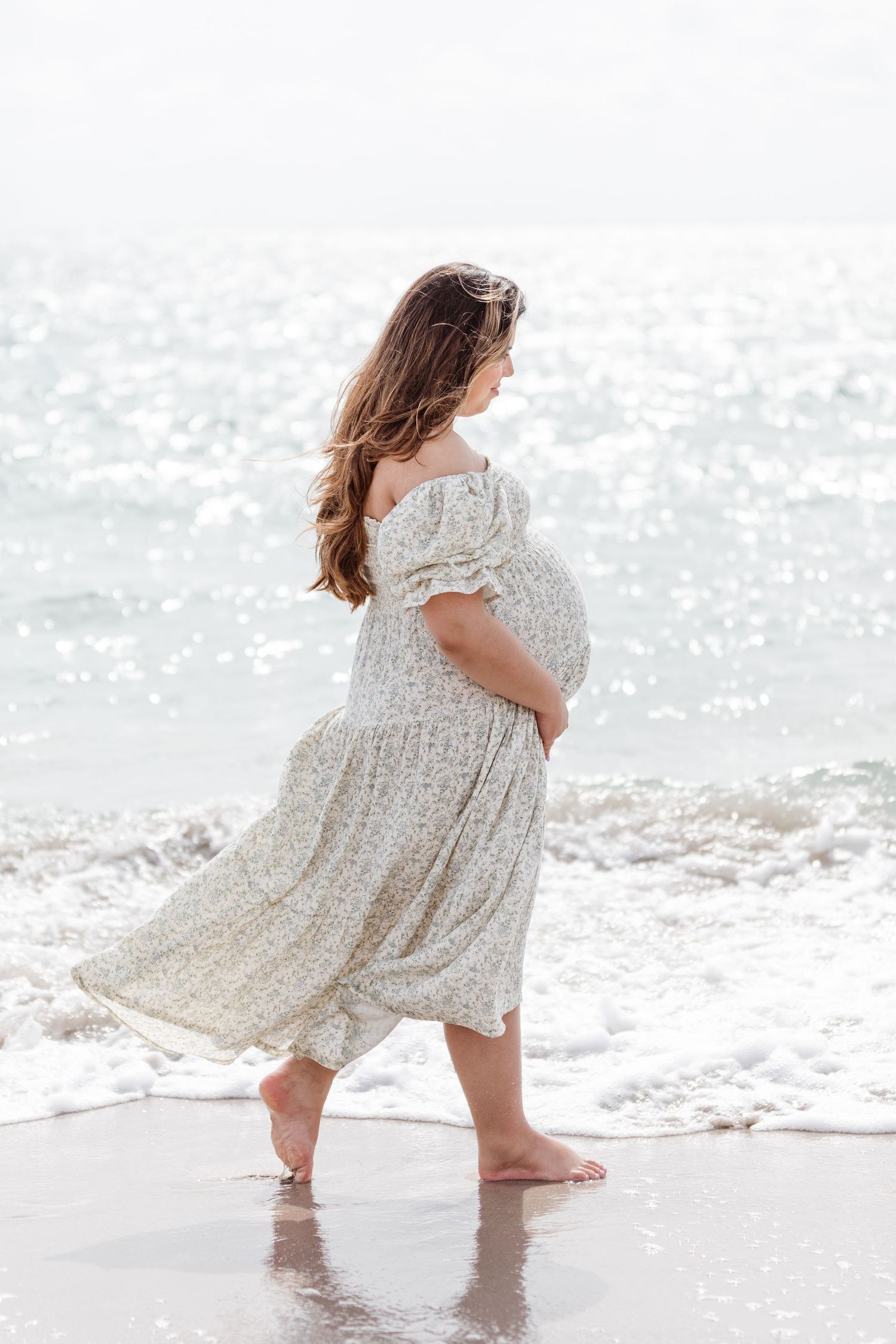 An expectant mother in a flowing white dress stands barefoot on a beach with waves lapping at her feet.