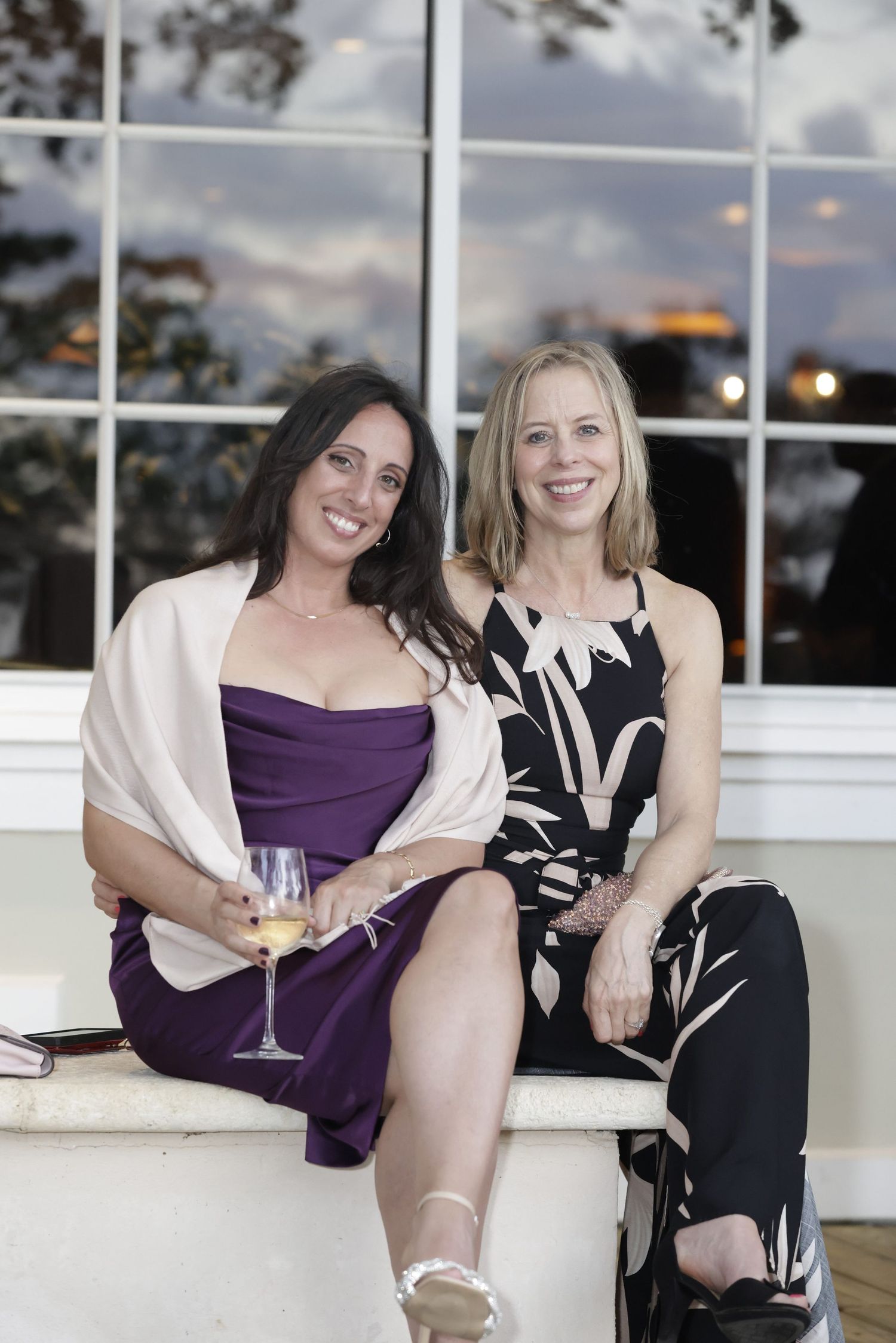 Two friends in purple and black dresses relax with drinks at an elegant outdoor venue during evening hours.