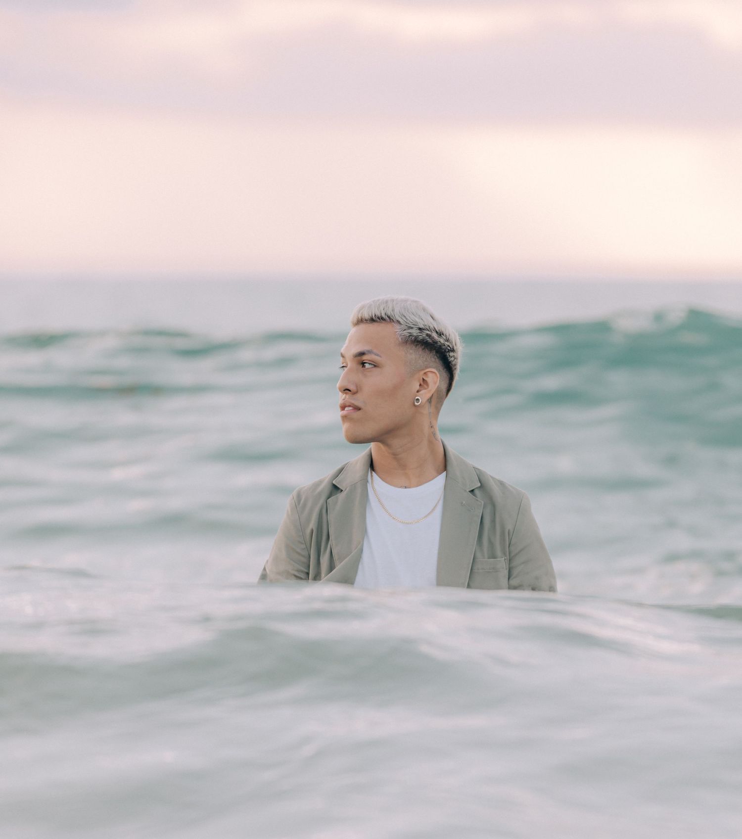 Person standing waist-deep in ocean waves against pink sky at sunset.
