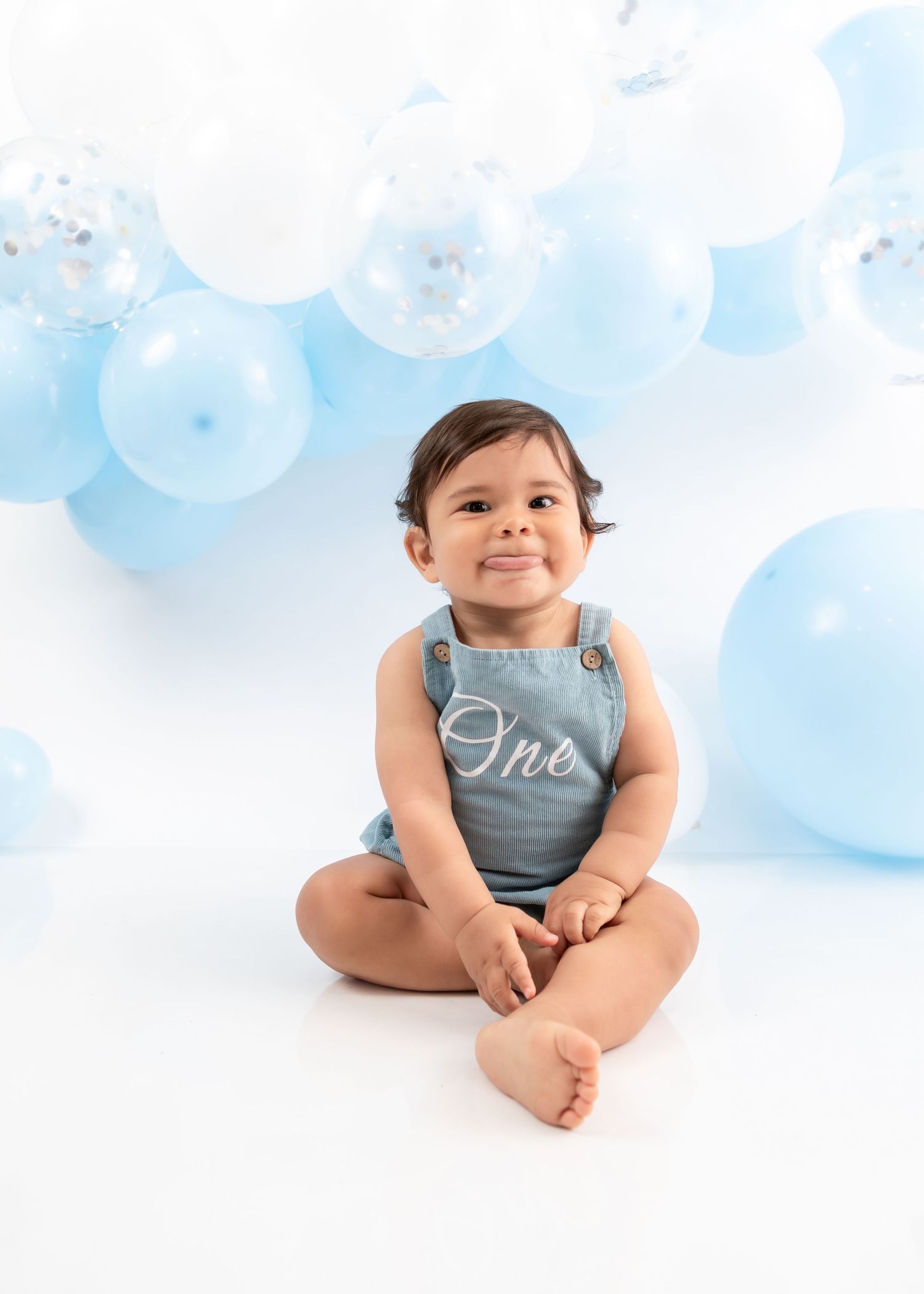 Baby in light blue romper sits against white backdrop with blue and white balloon decorations.