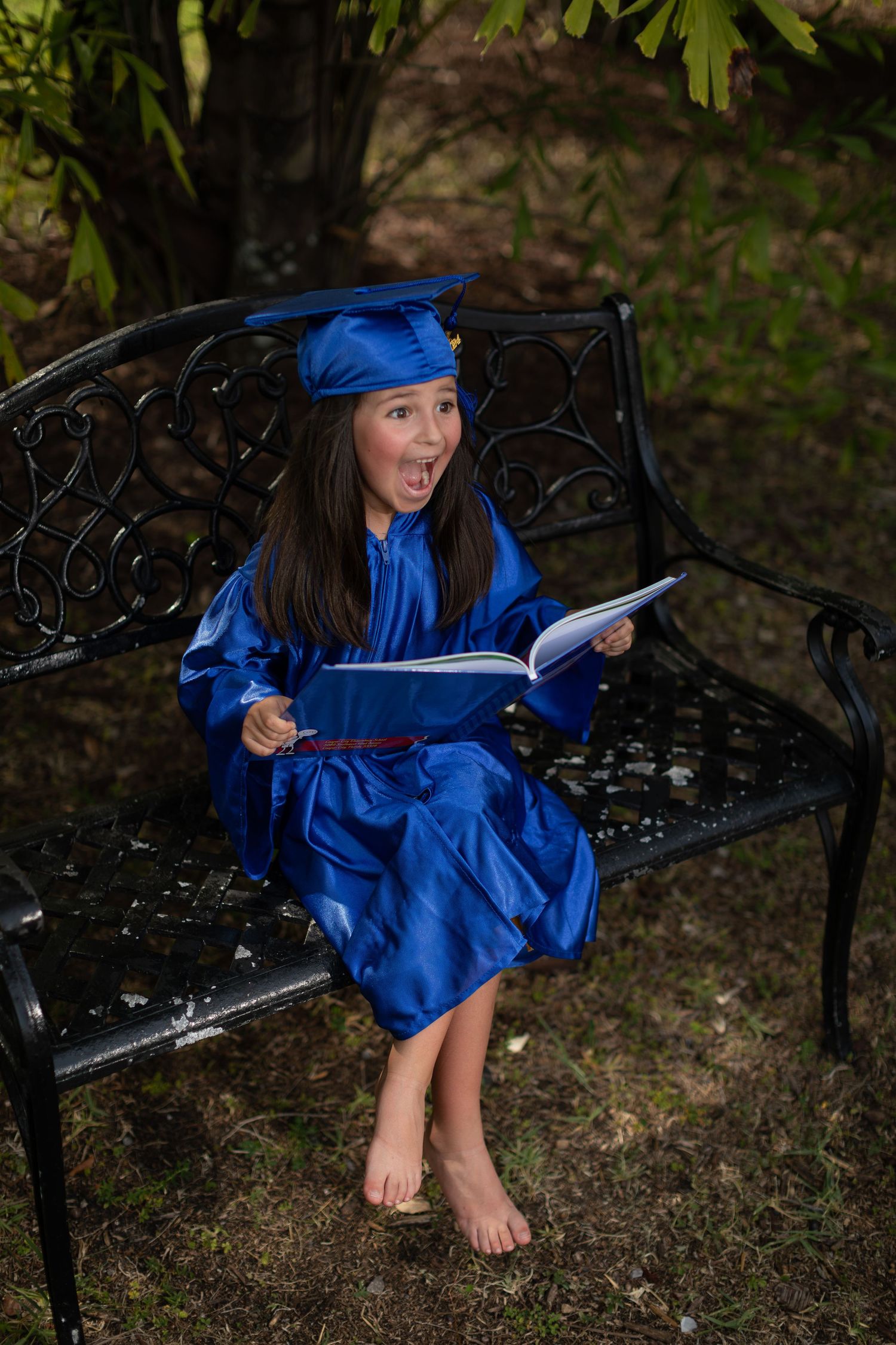 Graduate in blue cap and gown sits barefoot on decorative metal bench while holding a book with excited expression.