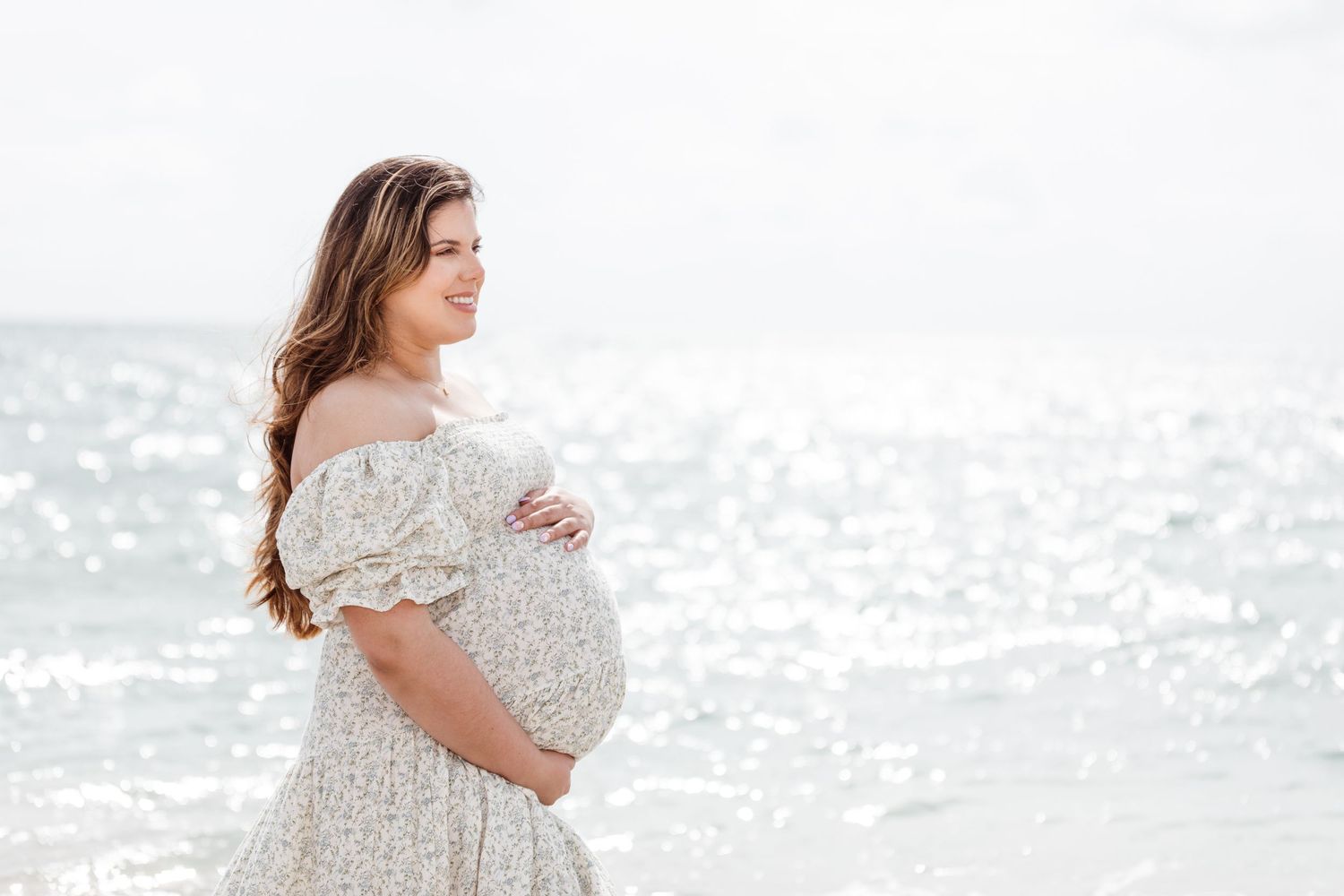 A pregnant woman in a lace dress stands by the ocean shore during a maternity photoshoot.