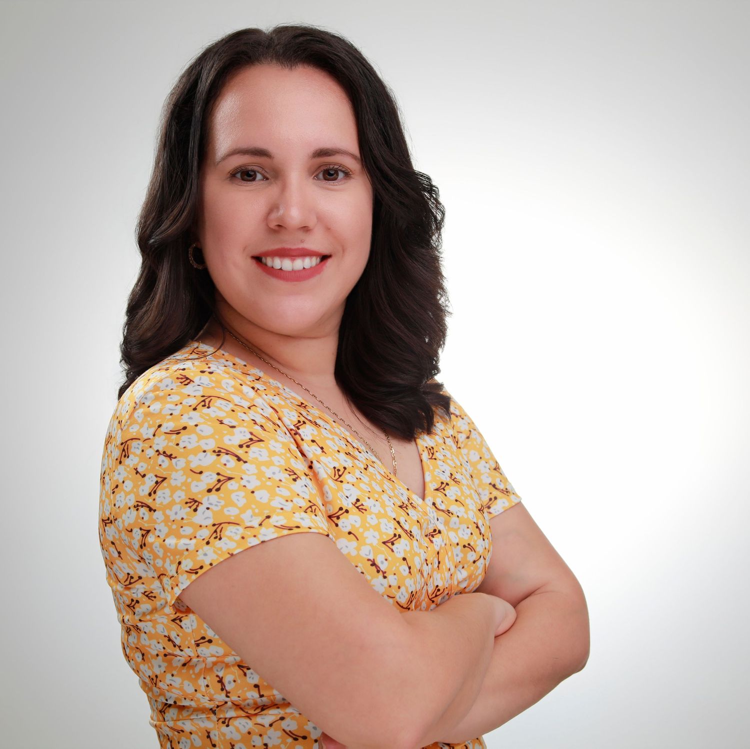 Friendly portrait in yellow floral top with wavy hair against white backdrop.