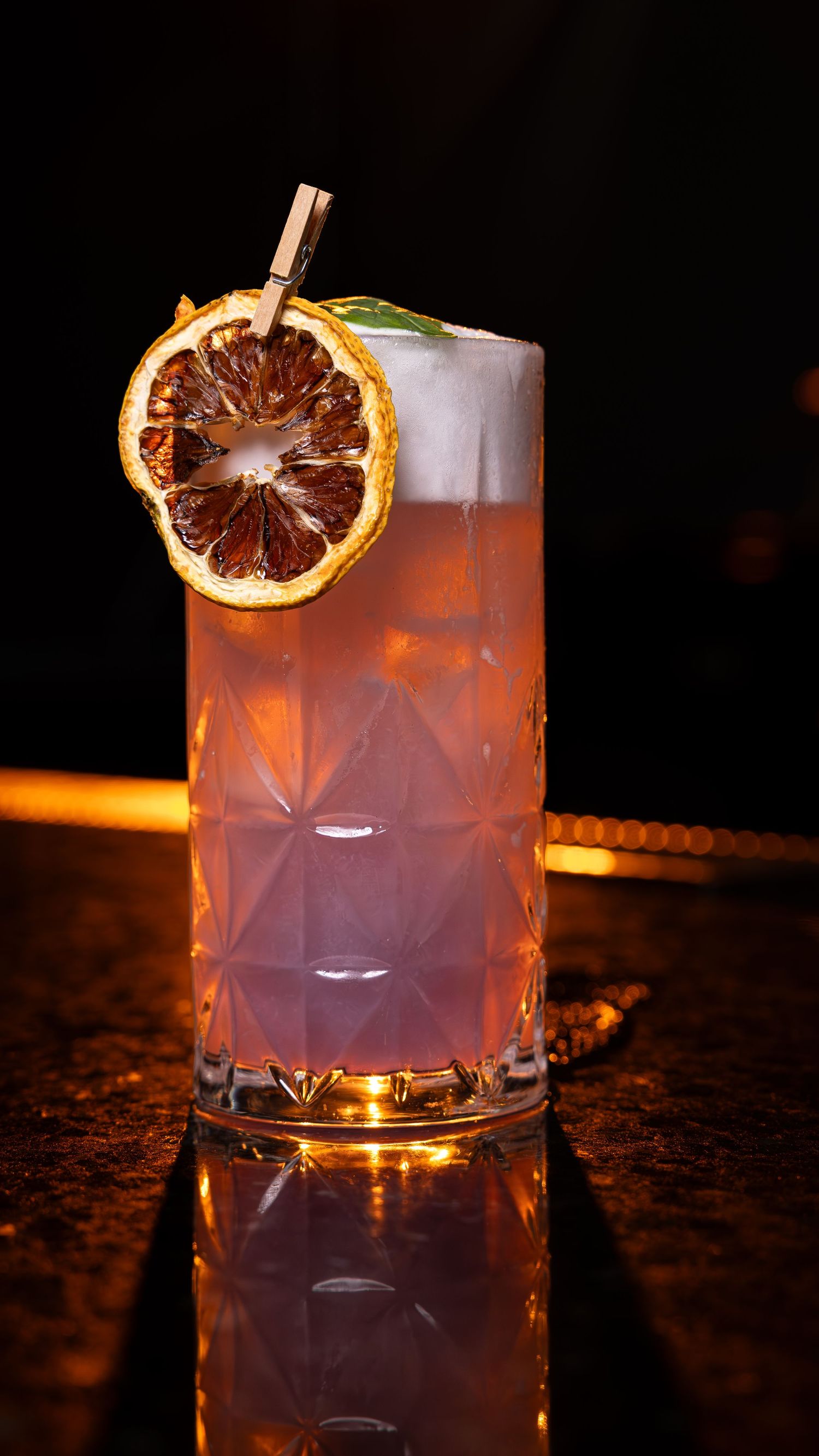 Craft cocktail in a rocks glass garnished with a dried citrus wheel and illuminated with warm ambient lighting.