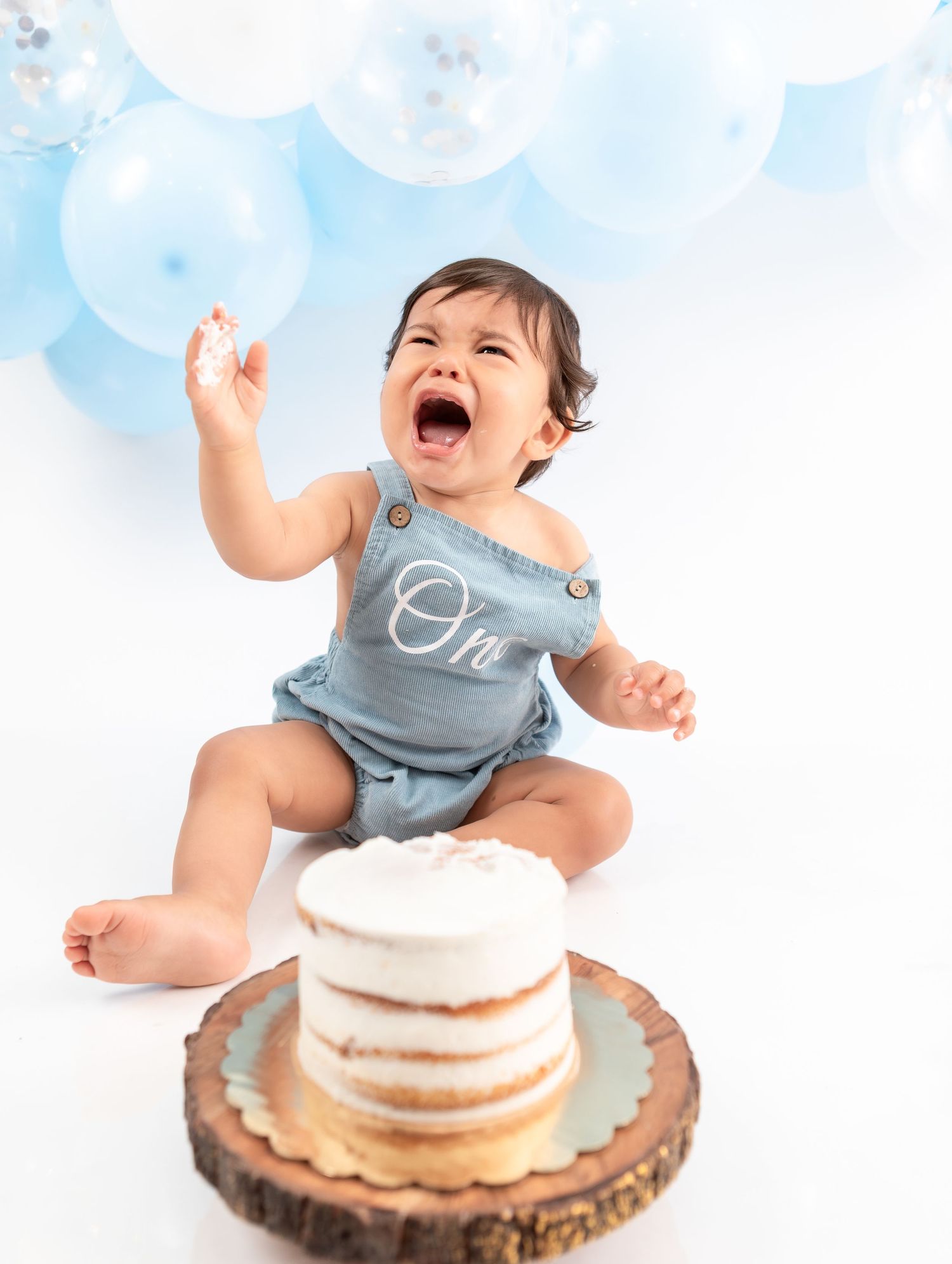 Excited baby in blue romper reaches up near white cake on wooden stand with balloon backdrop.