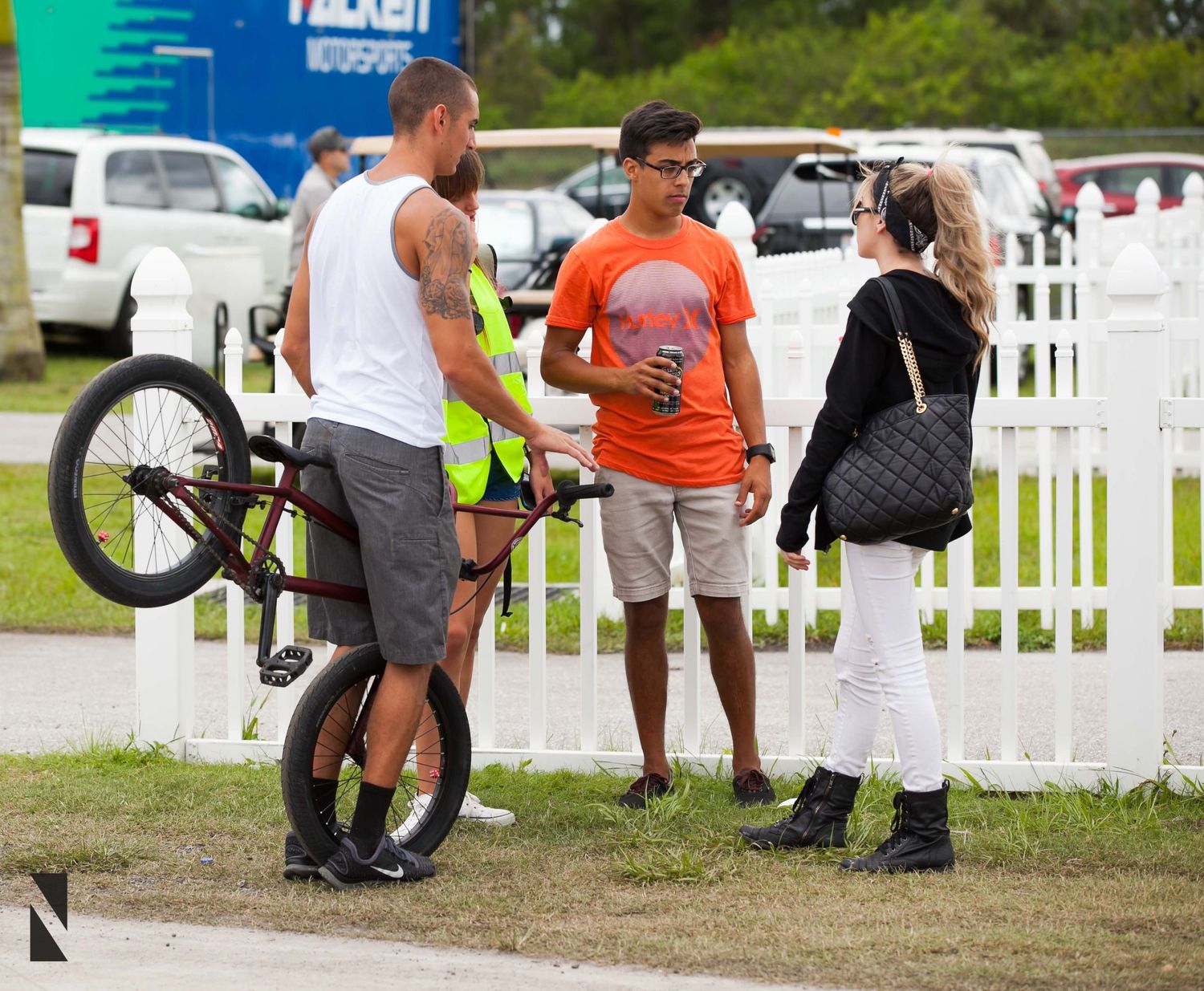 People gather outdoors near a white picket fence as someone holds a BMX bicycle at a sports event.