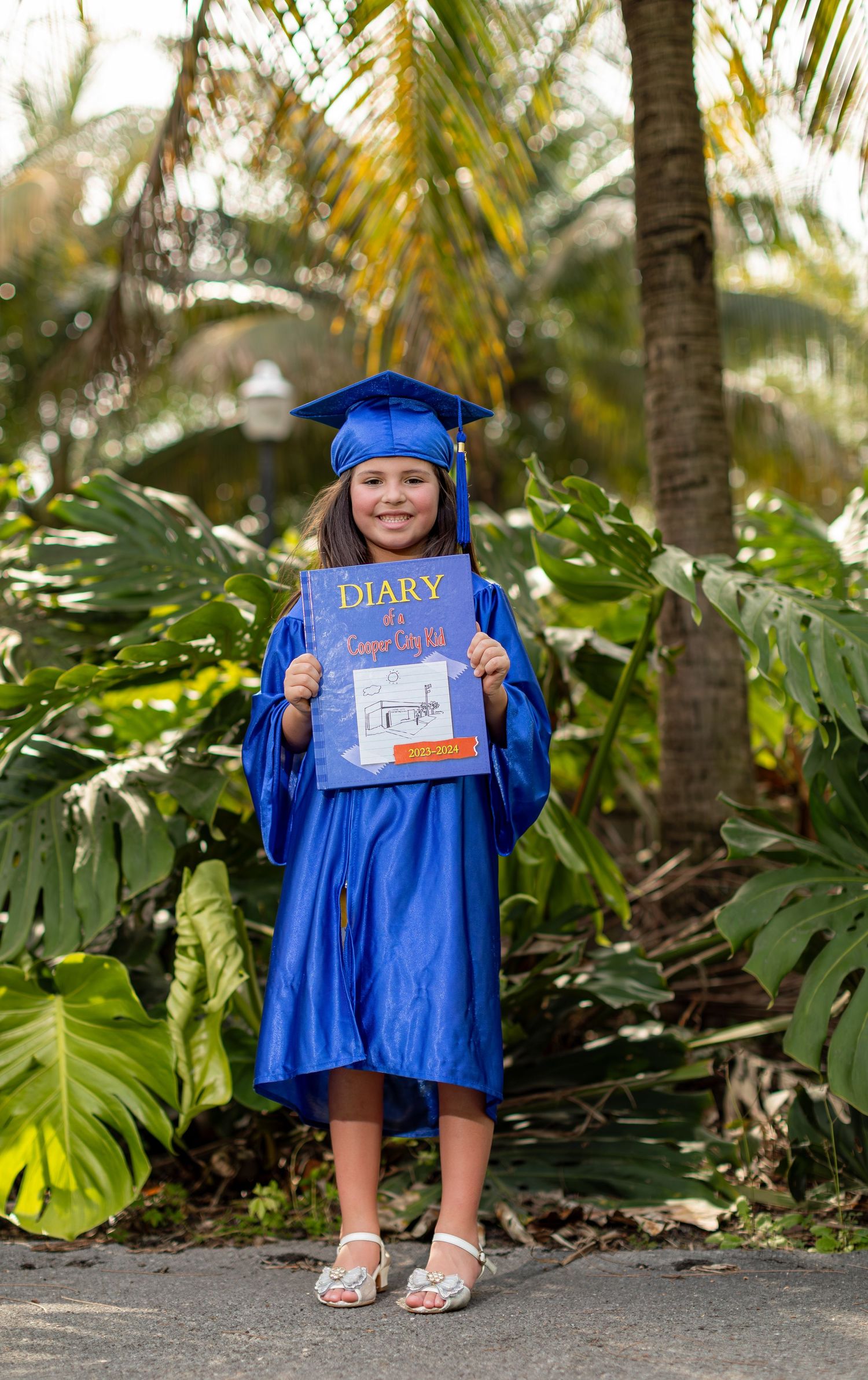 A graduate in bright blue cap and gown holds up a Diary of a Wimpy Kid book while standing among tropical palm trees.