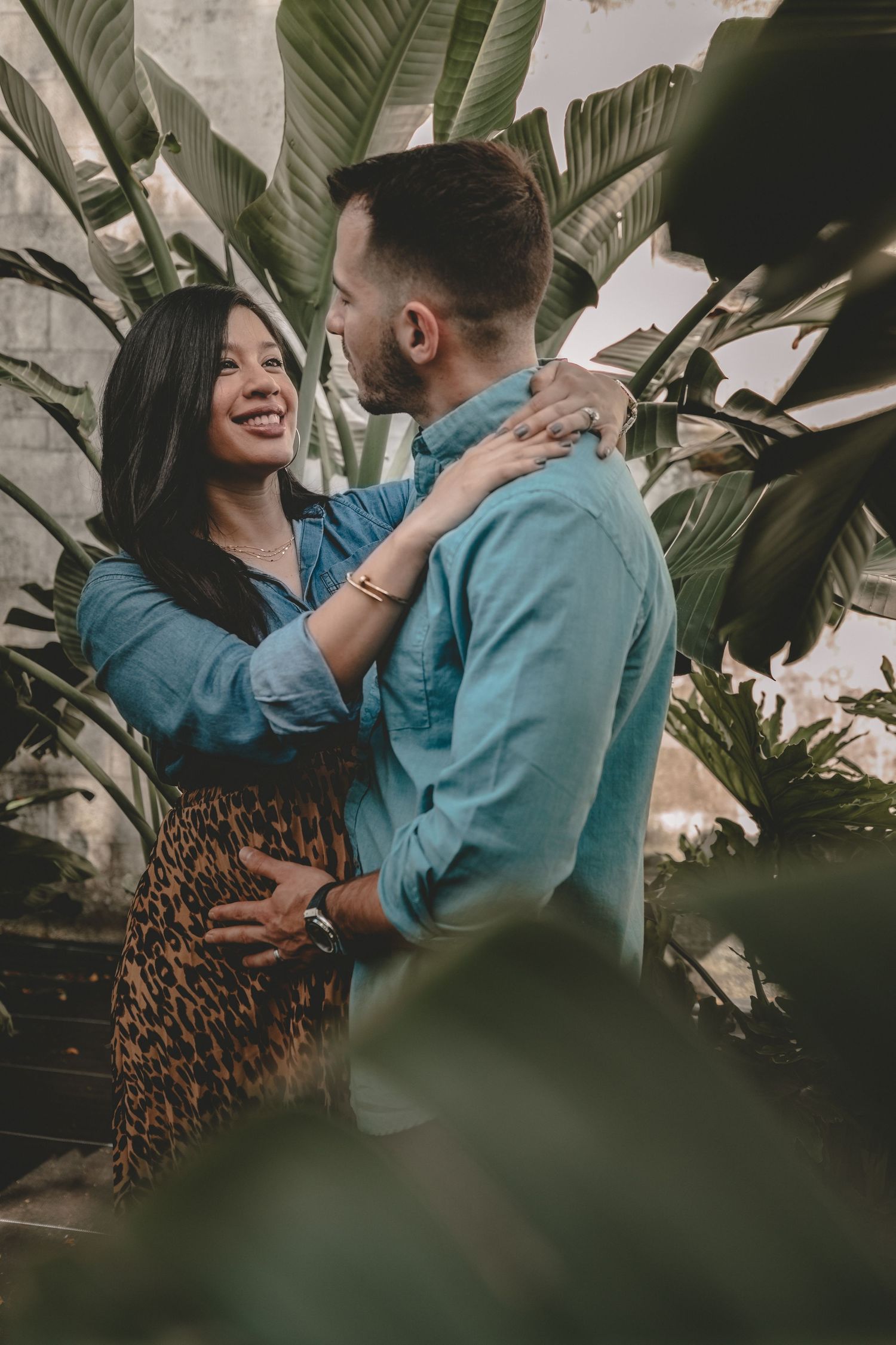 A romantic couple embraces among tropical plants while wearing coordinated blue outfits.