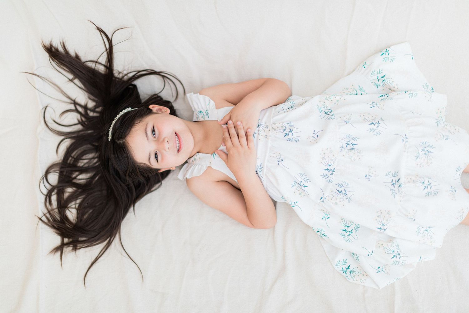A child lies on white floor with dark hair spread out, wearing a floral sundress.