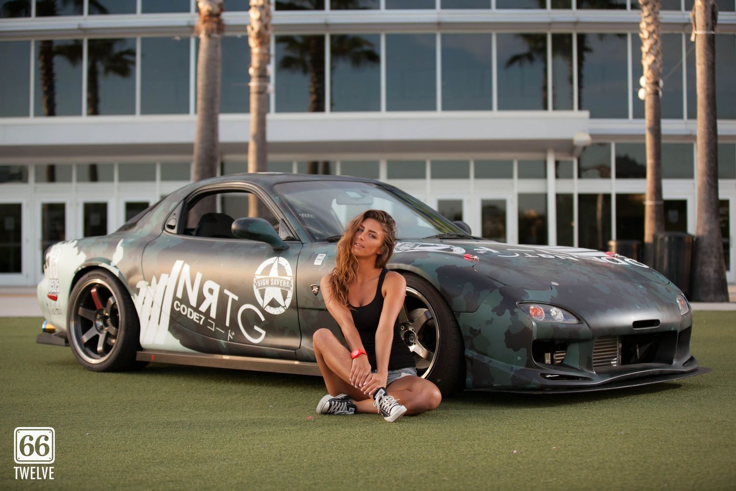Custom black sports car with bold graphics parked on grass lawn with palm trees and building in background.