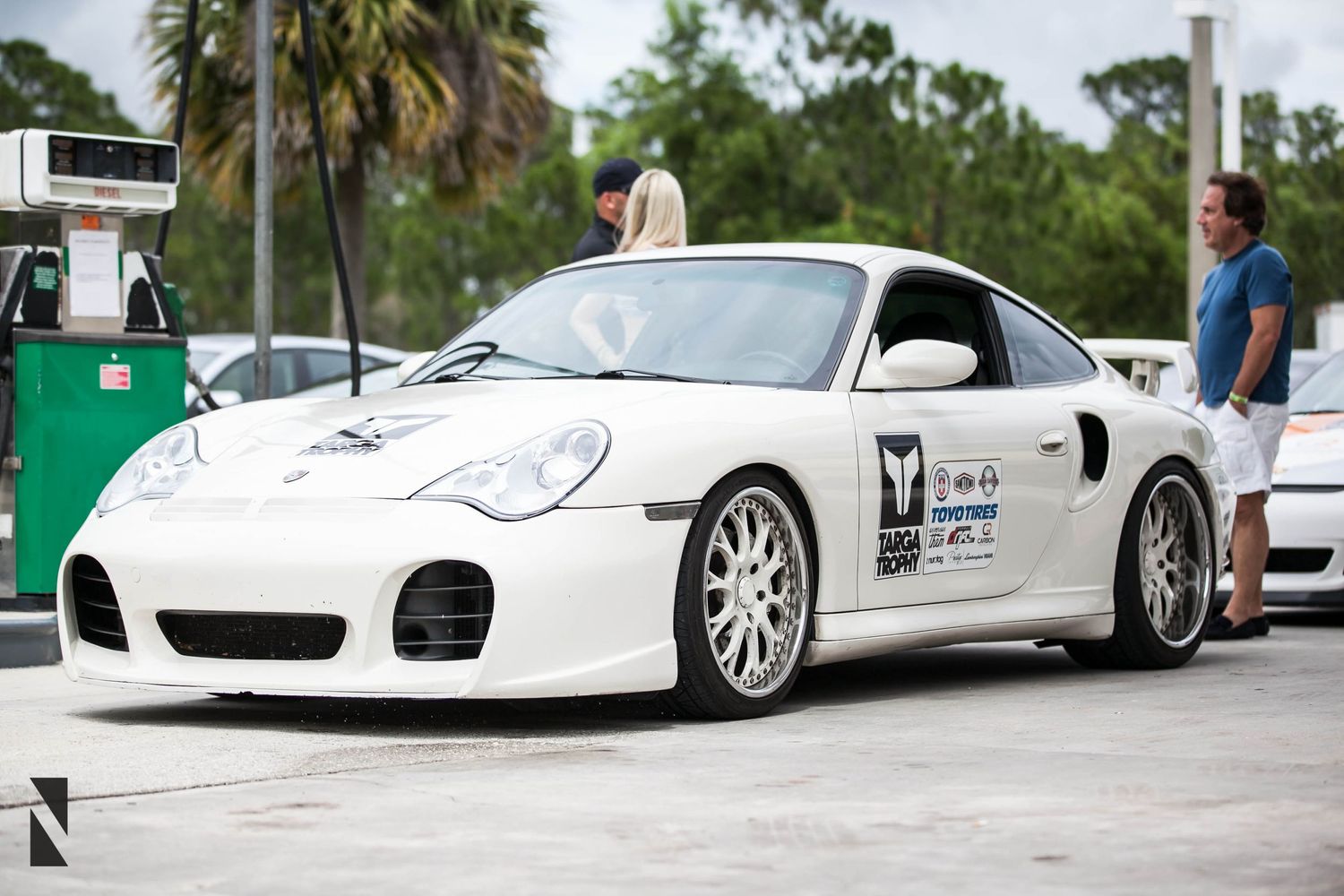 White Porsche 911 Turbo sports car with racing decals parked at an outdoor event with palm trees in background.