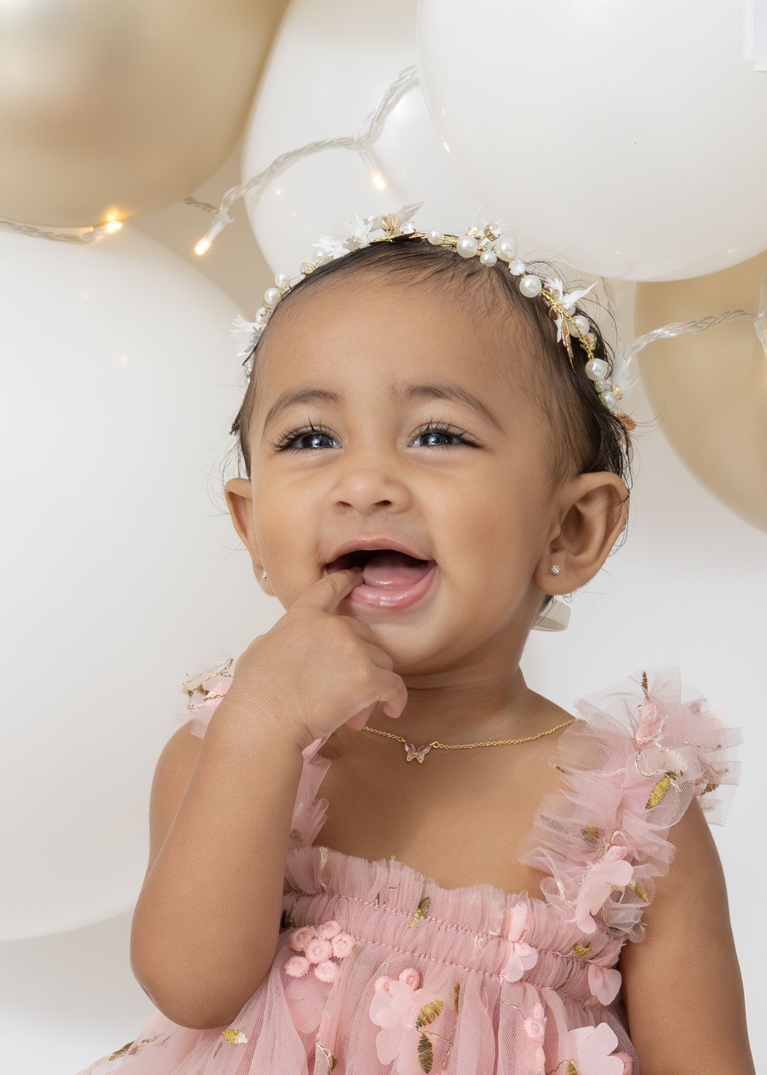 A smiling child wearing a pink tulle dress with a floral headpiece poses against a light background with balloons.