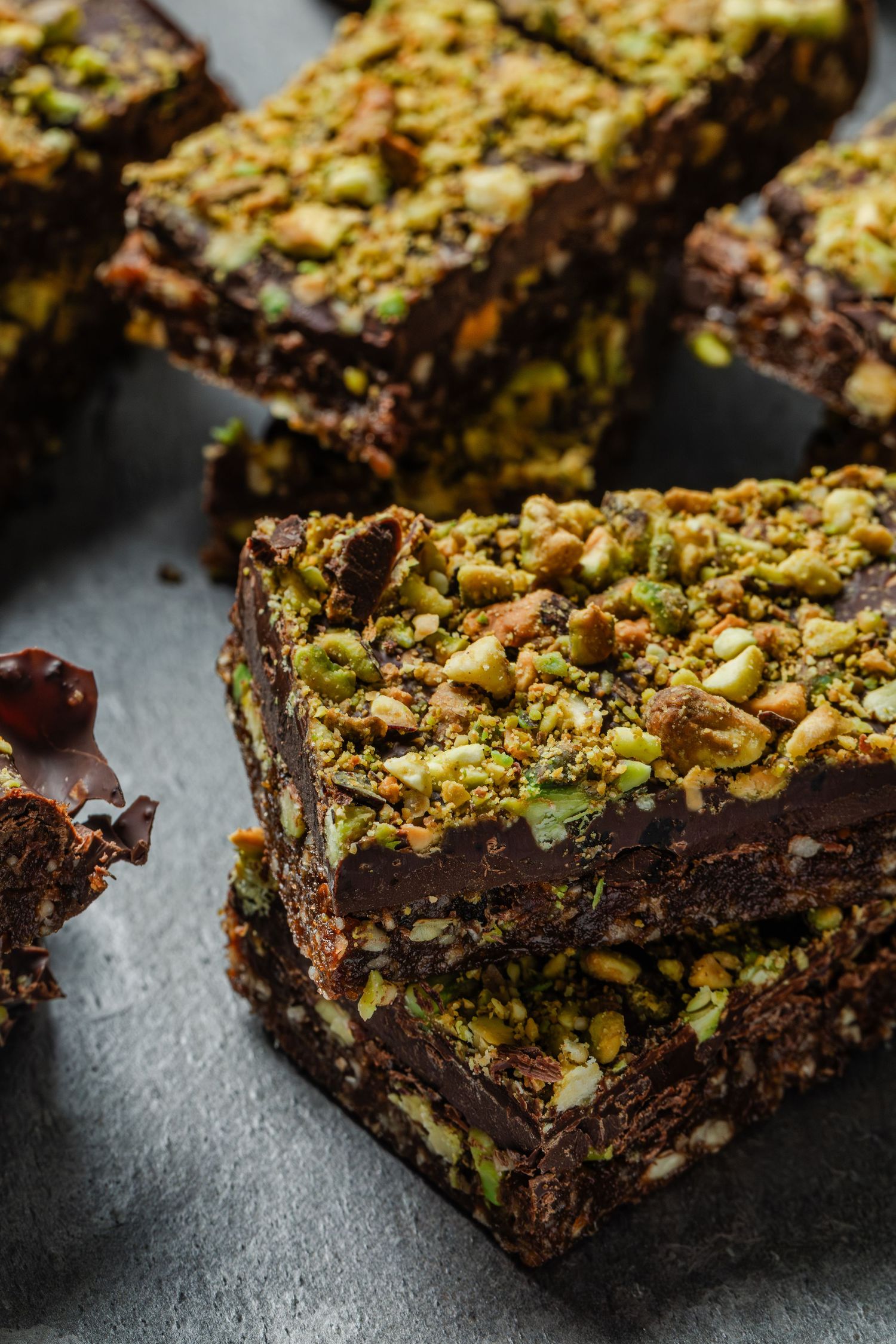 Close-up shot of dark chocolate brownies topped with crushed pistachios on a grey surface in warm lighting.