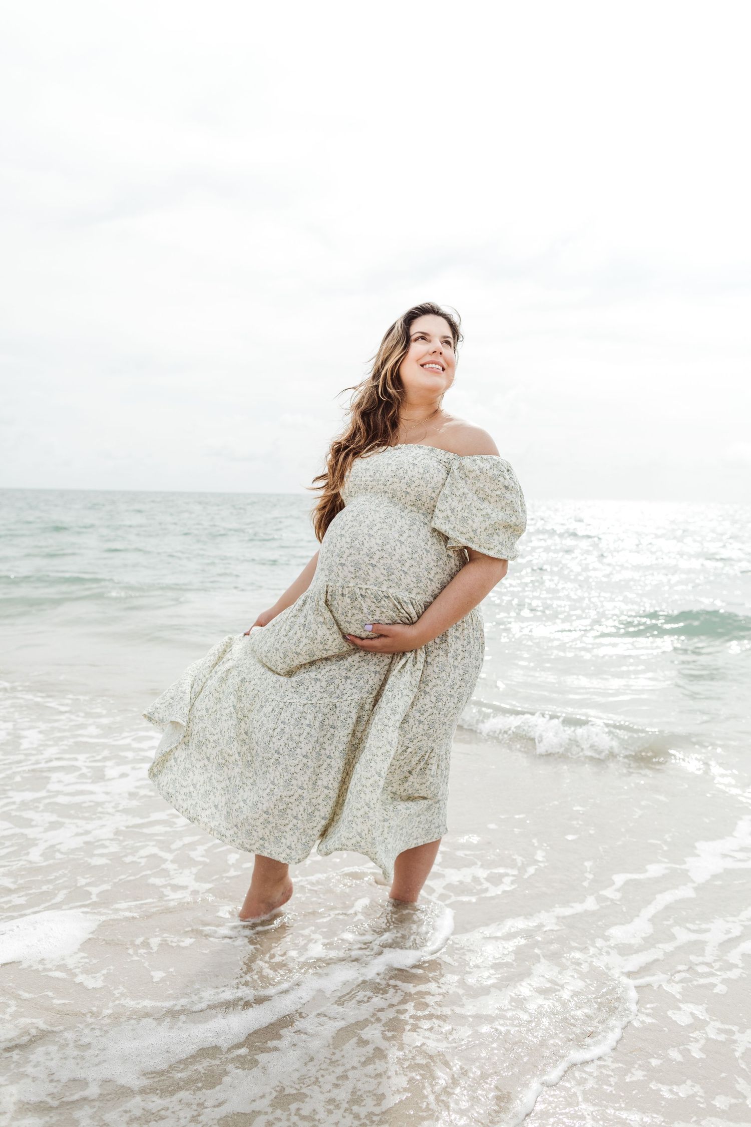 Expectant woman in floral dress stands in ocean waves at beach during maternity photoshoot.