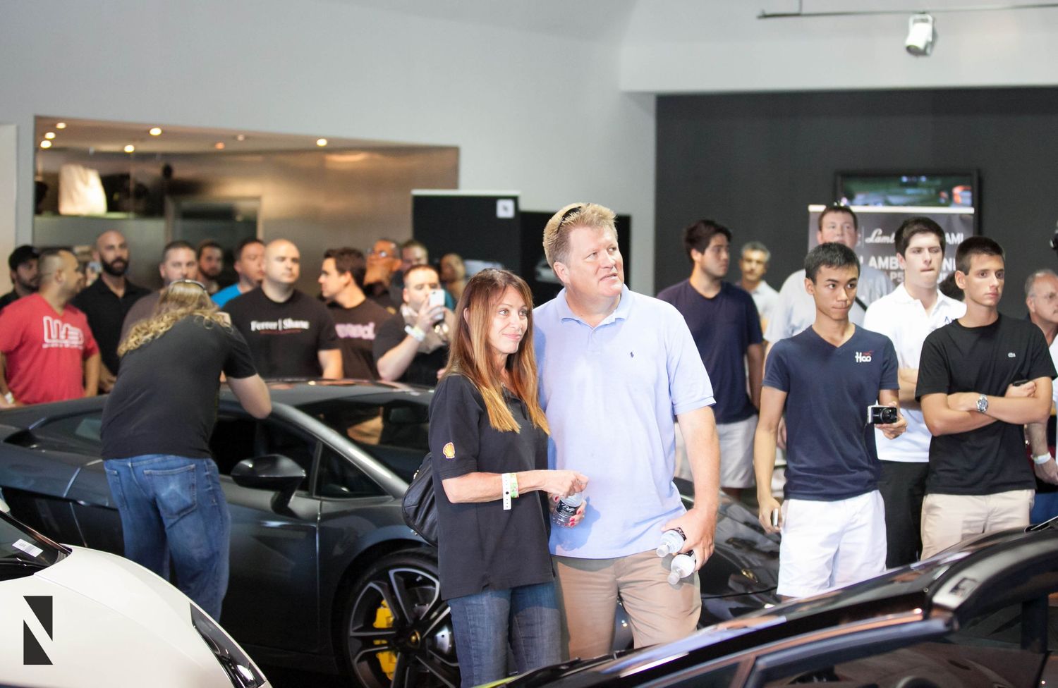 A crowd of people gathered around cars at an indoor automotive event or showroom.