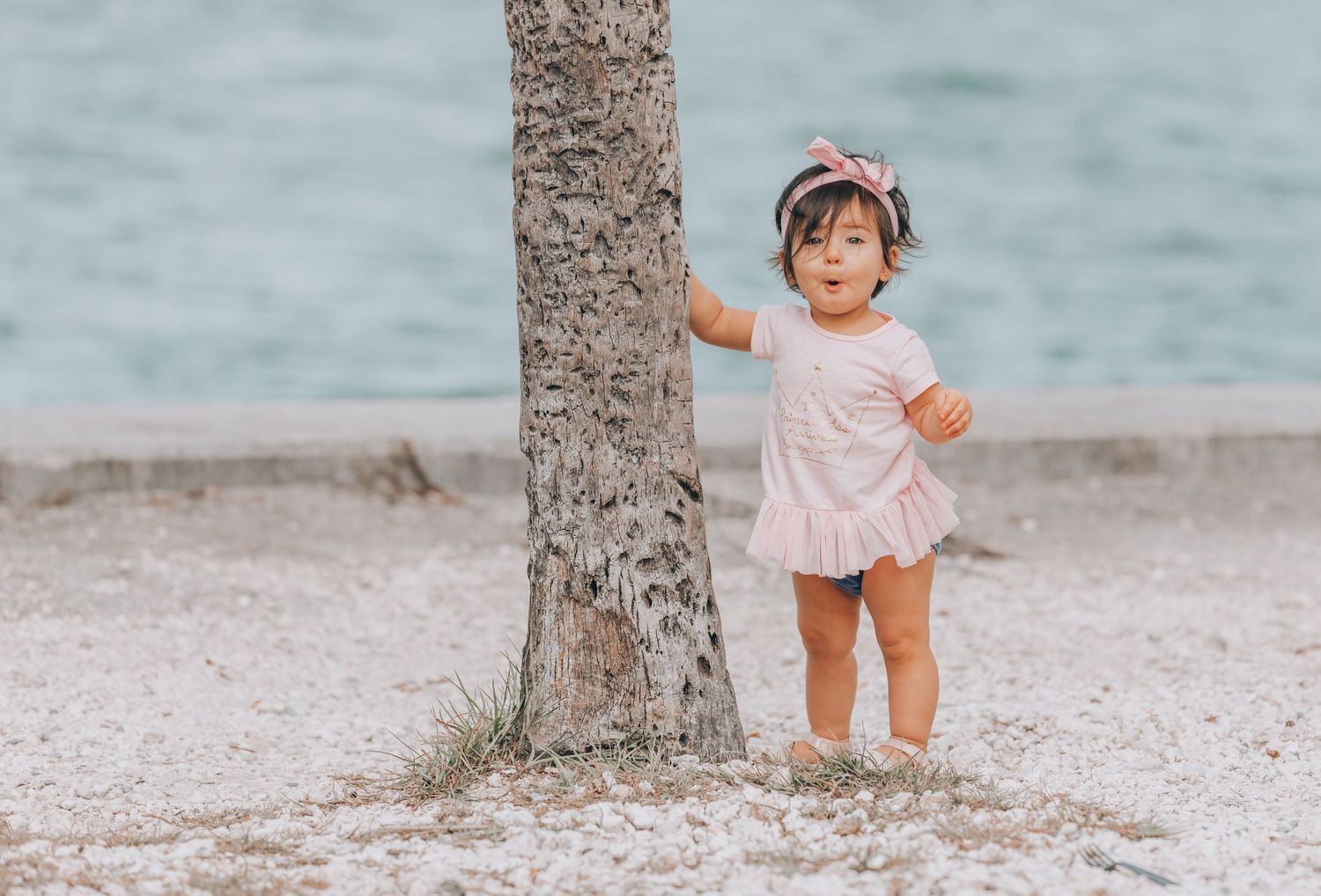 Small child wearing a pink outfit and headband stands next to a palm tree trunk on a tropical beach by the water.