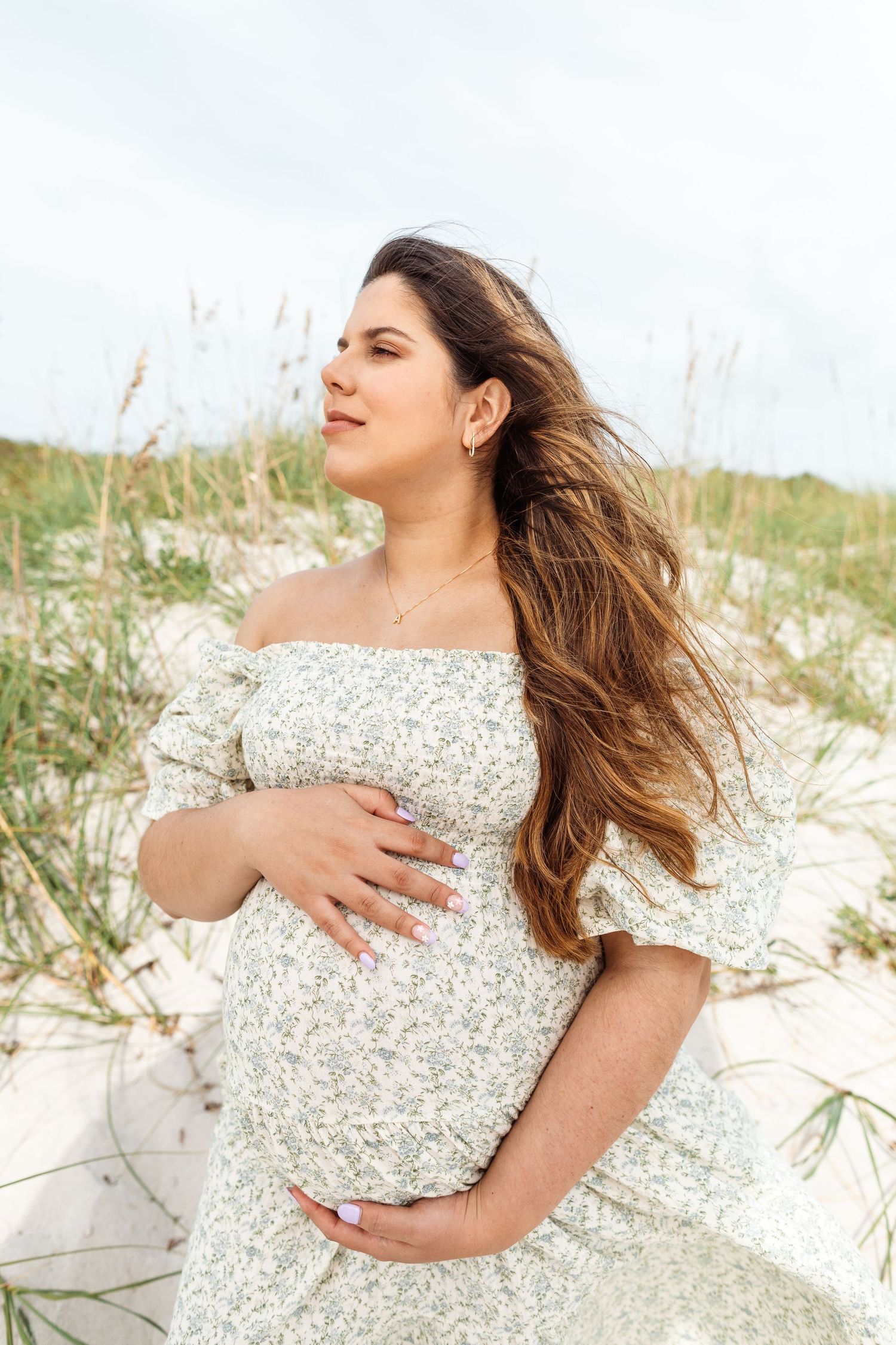 Pregnant woman in flowing white dress poses serenely on beach sand dunes with windswept hair.