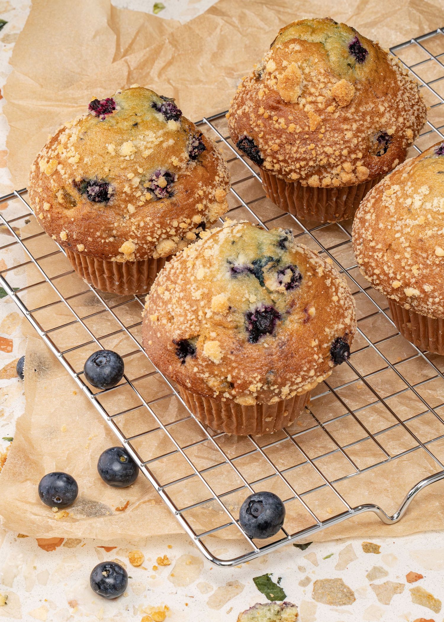 Fresh blueberry muffins cooling on a wire rack with crumb topping and scattered fresh berries on brown paper.