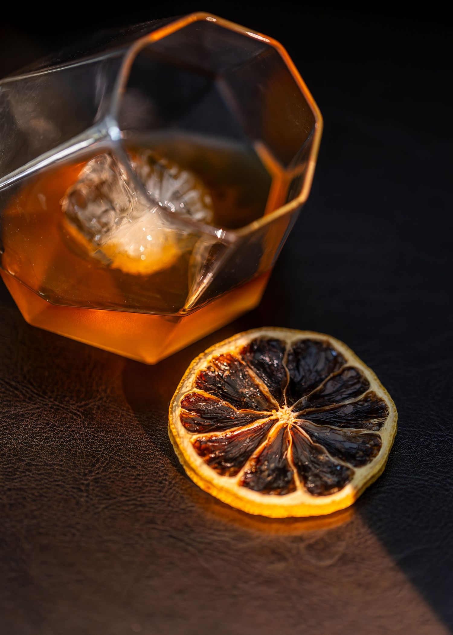 Glass filled with amber liquid and garnished with a dried citrus wheel catches warm light in dramatic close-up shot.