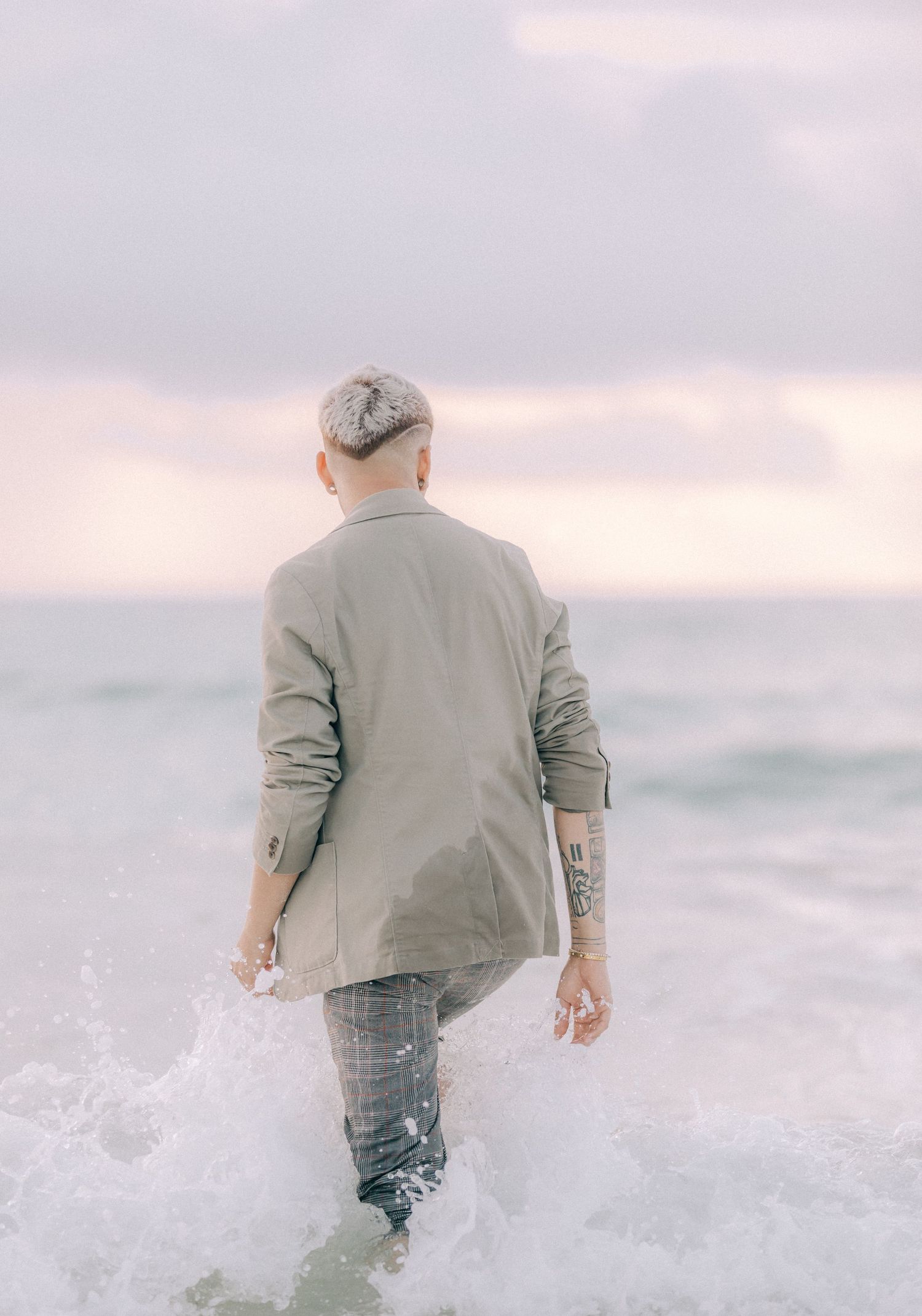 Person in gray blazer walking through ocean waves at sunset.