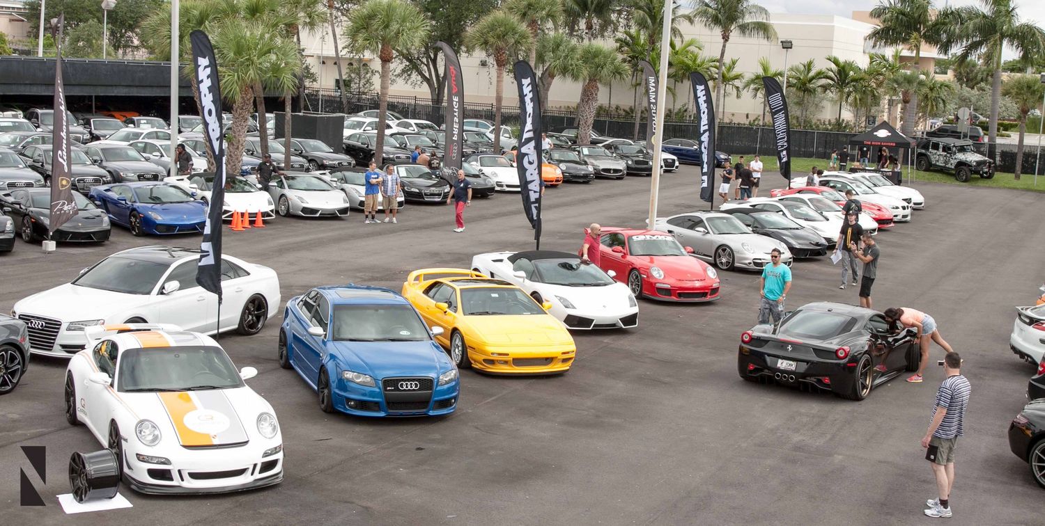 A collection of luxury sports cars including Porsche, Ferrari and other high-end vehicles parked in an outdoor lot.