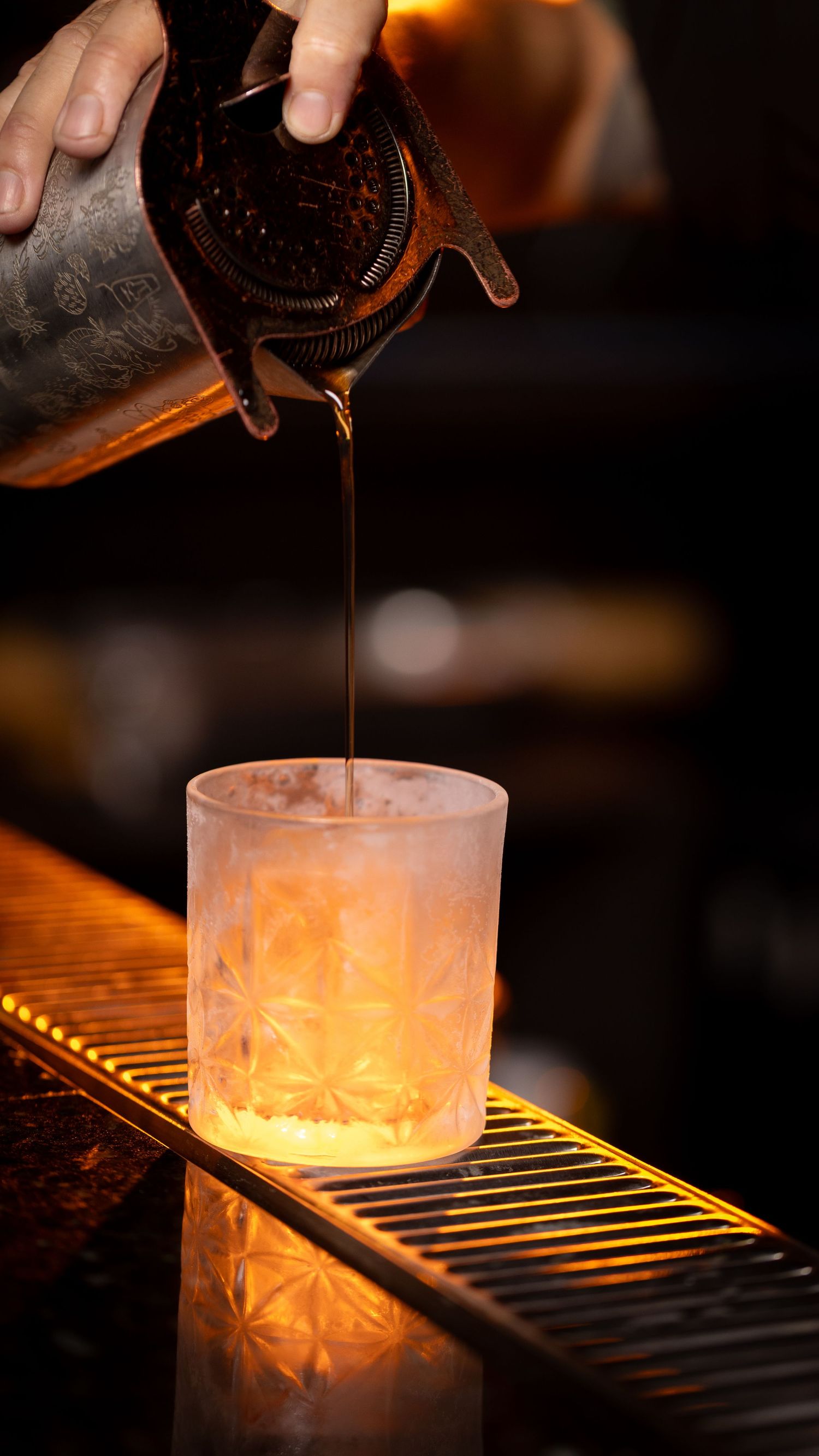 A bartender pours a dark cocktail into a frosted glass on an illuminated bar counter, creating a warm ambiance.