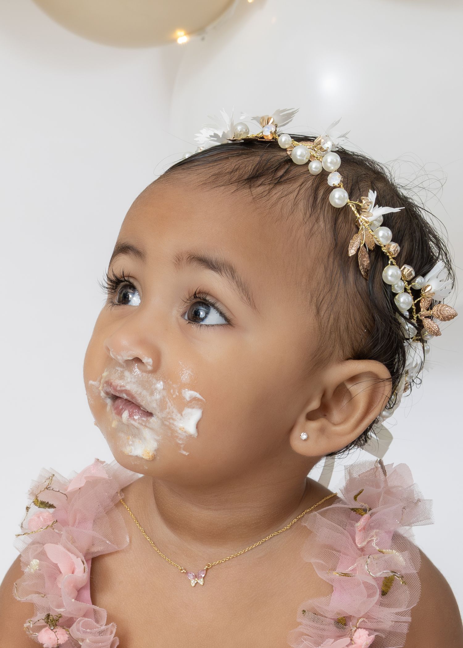 A baby wearing a pearl floral headband and pink tulle dress has cake frosting smeared on their face during a cake smash photo.