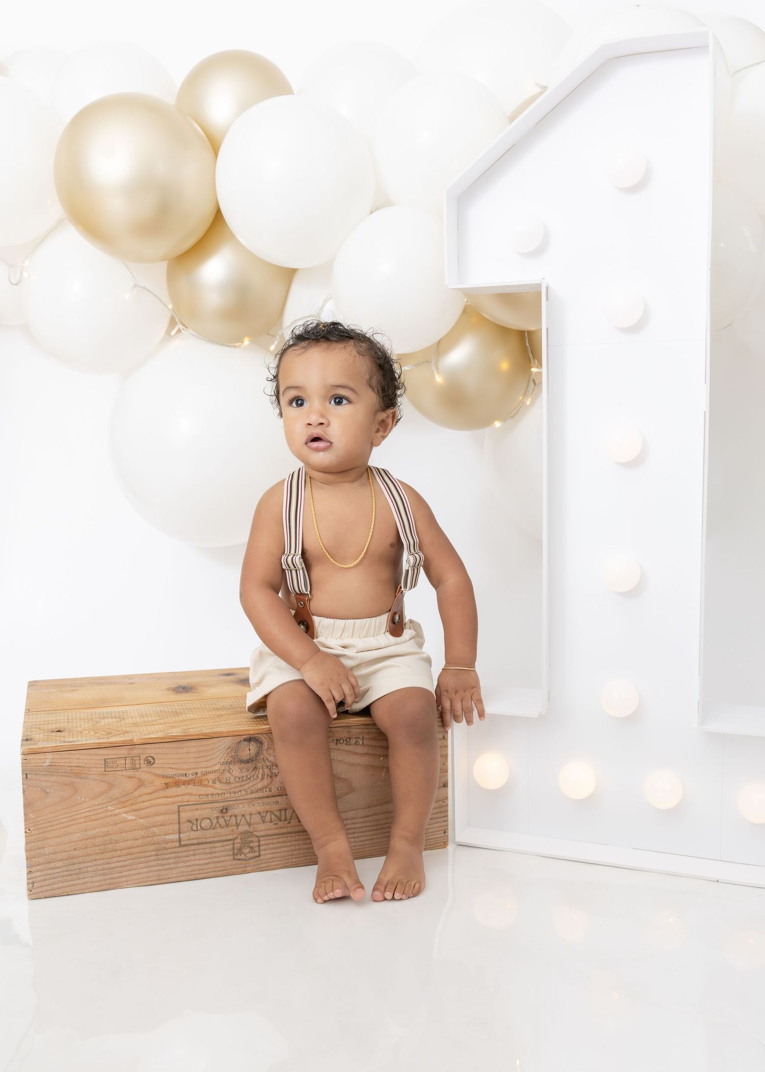 A baby in white overalls poses against a backdrop with gold and white balloons and a wooden crate.