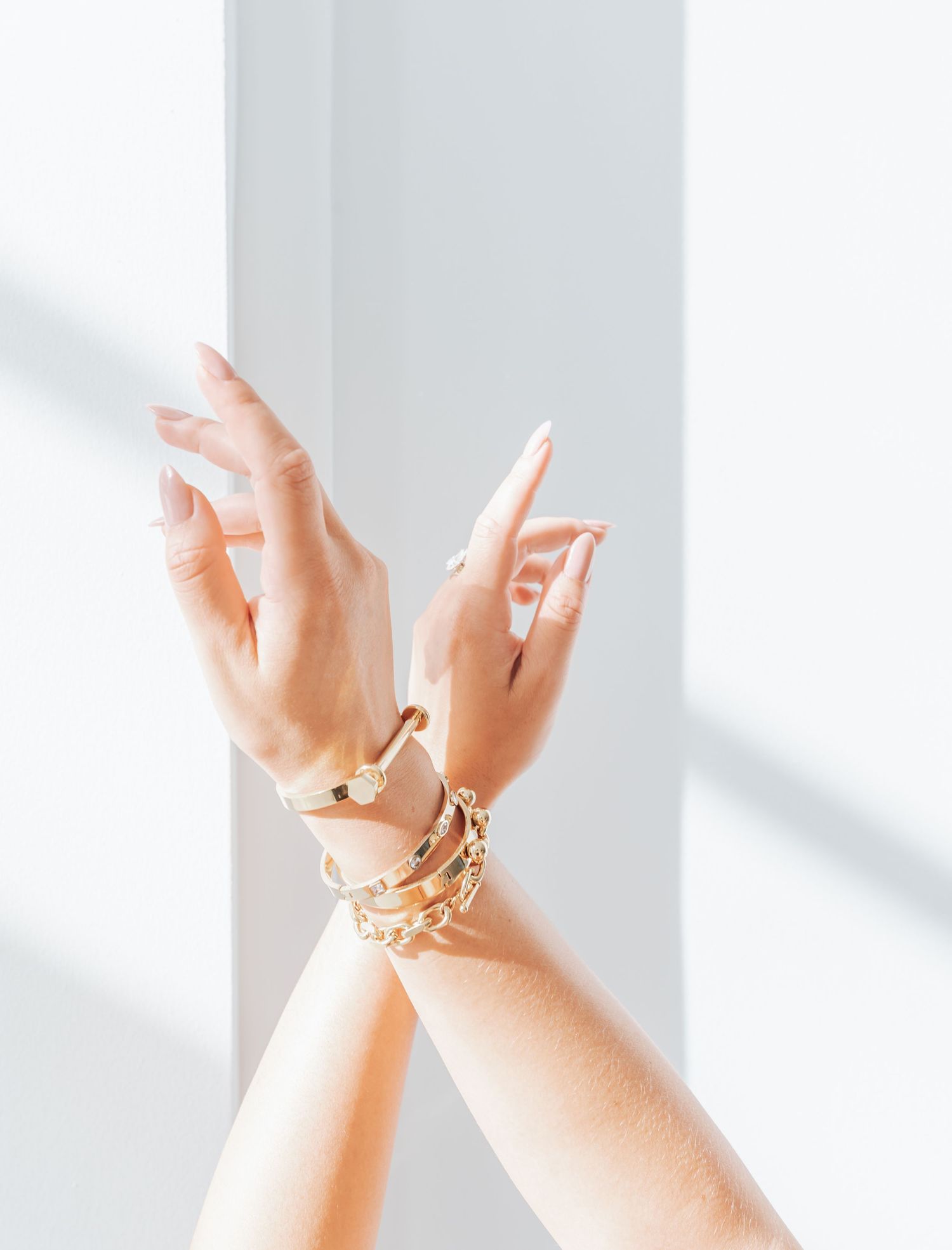 Crossed hands with gold bracelets and rings against white background with shadows.