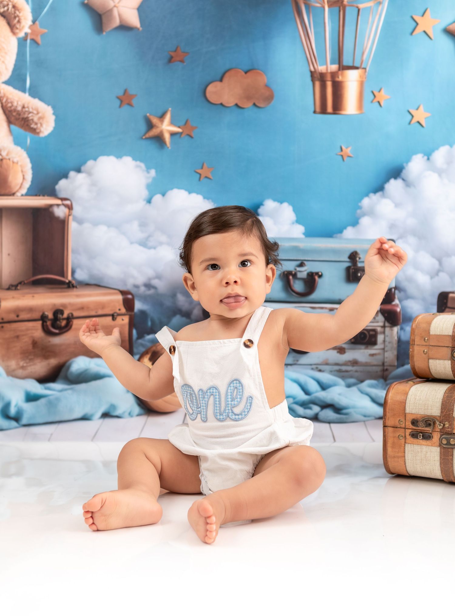 Baby in white romper sits among travel-themed decor with blue sky and cloud background.
