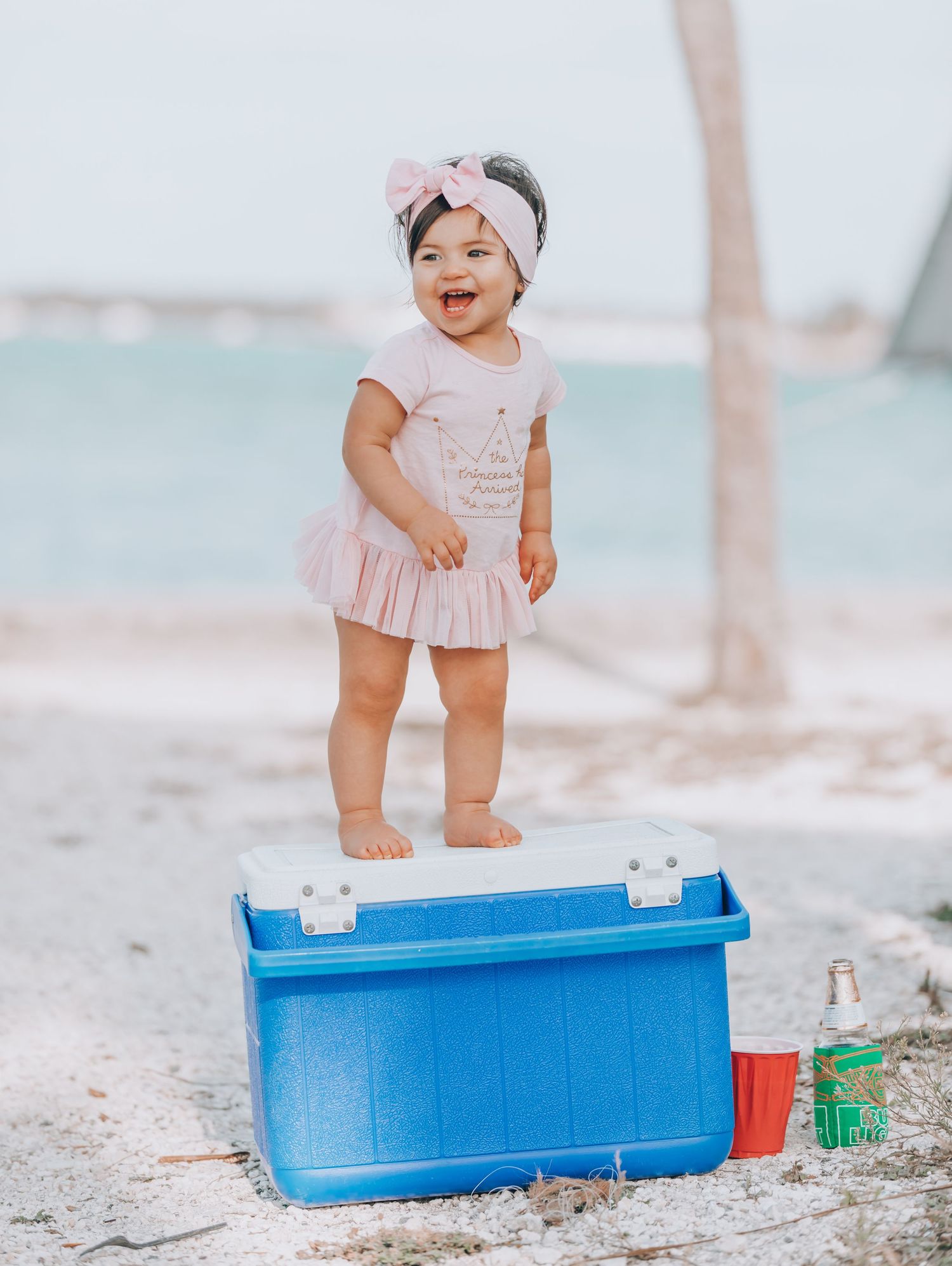 A smiling toddler in a pink dress and headband stands on a beach next to a blue cooler and drinks on the sand.