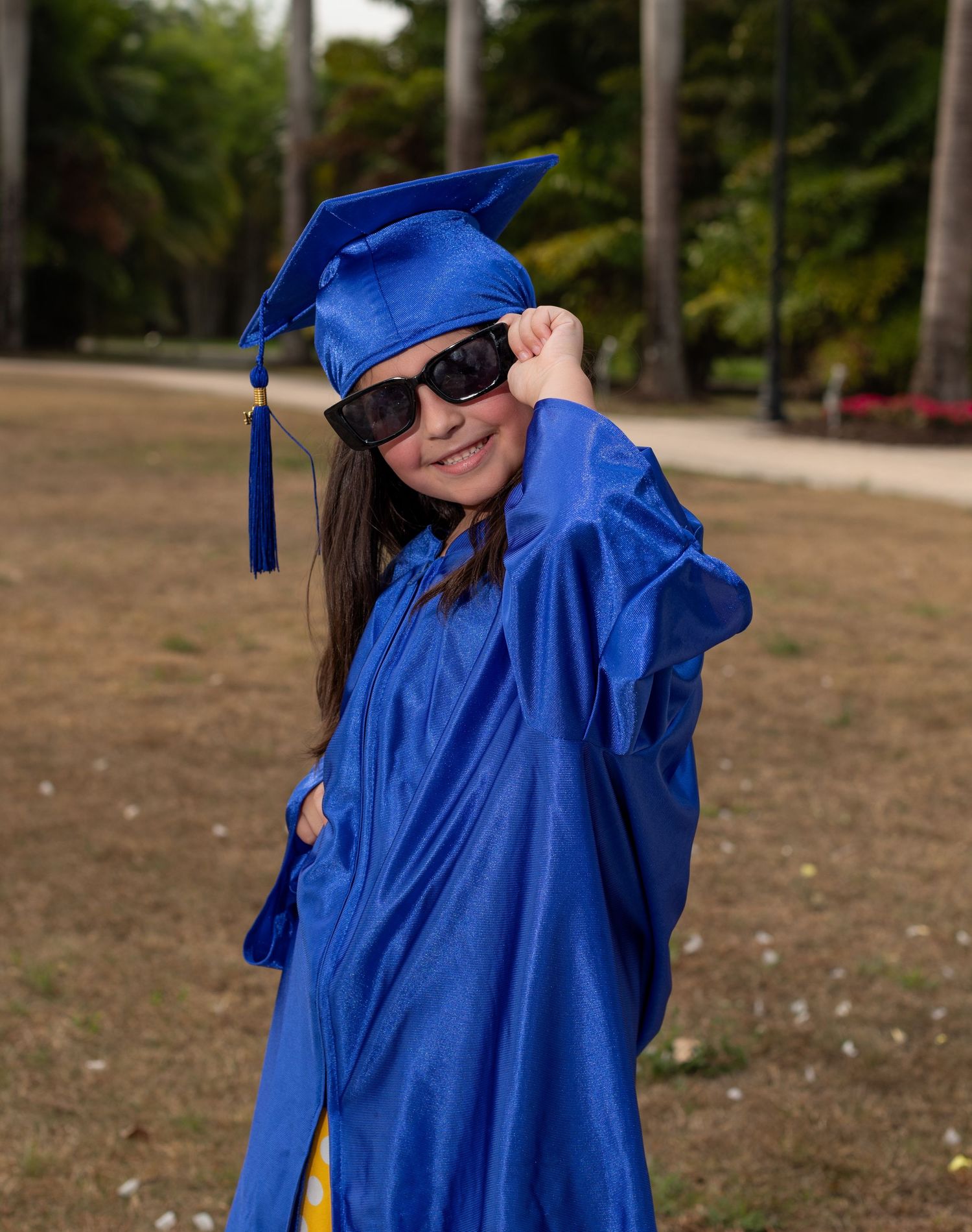 Graduate in bright blue cap and gown poses outdoors wearing sunglasses against palm tree background at graduation ceremony.