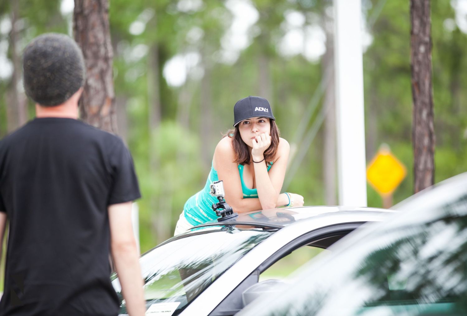 Woman in blue top leans against white car in outdoor setting with pine trees in background.